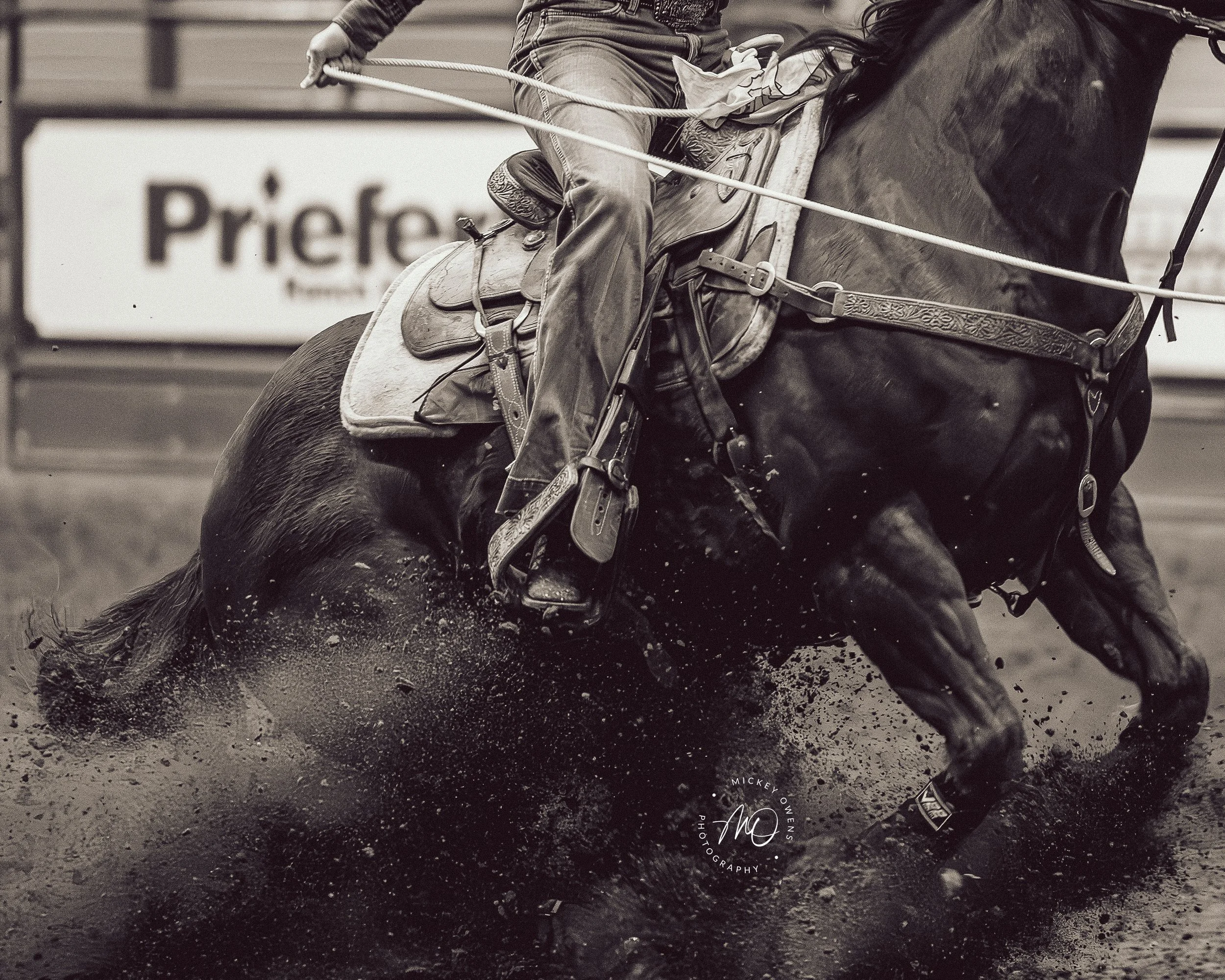 A person riding a horse at a gallop on a dirt track, with motion splash and dirt flying beneath the horse's hooves. The rider is holding the reins and leaning slightly forward.