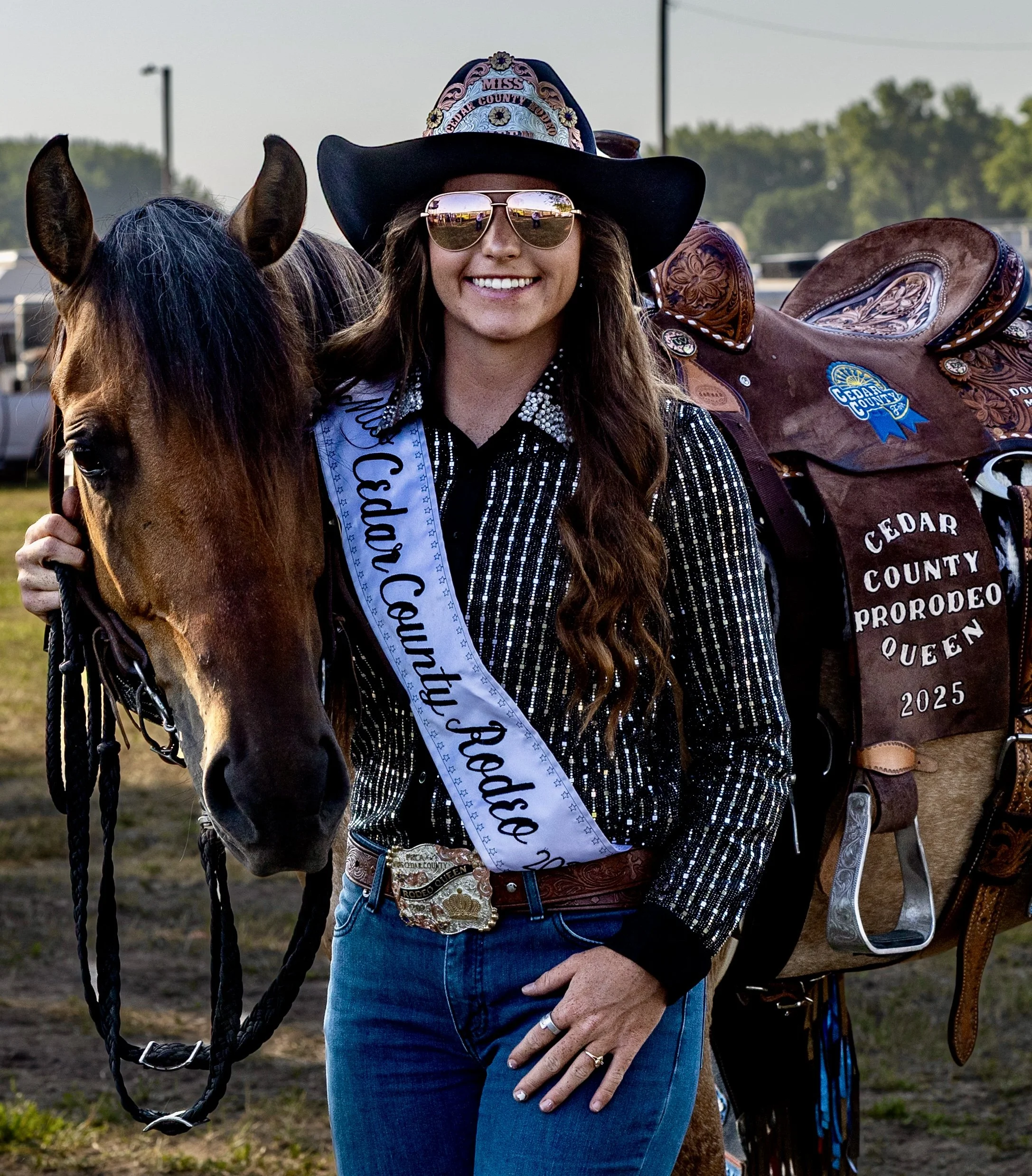 Woman wearing a cowboy hat, sunglasses, and a sash, standing beside a horse at an outdoor rodeo event. Sky and trees are visible in the background.
