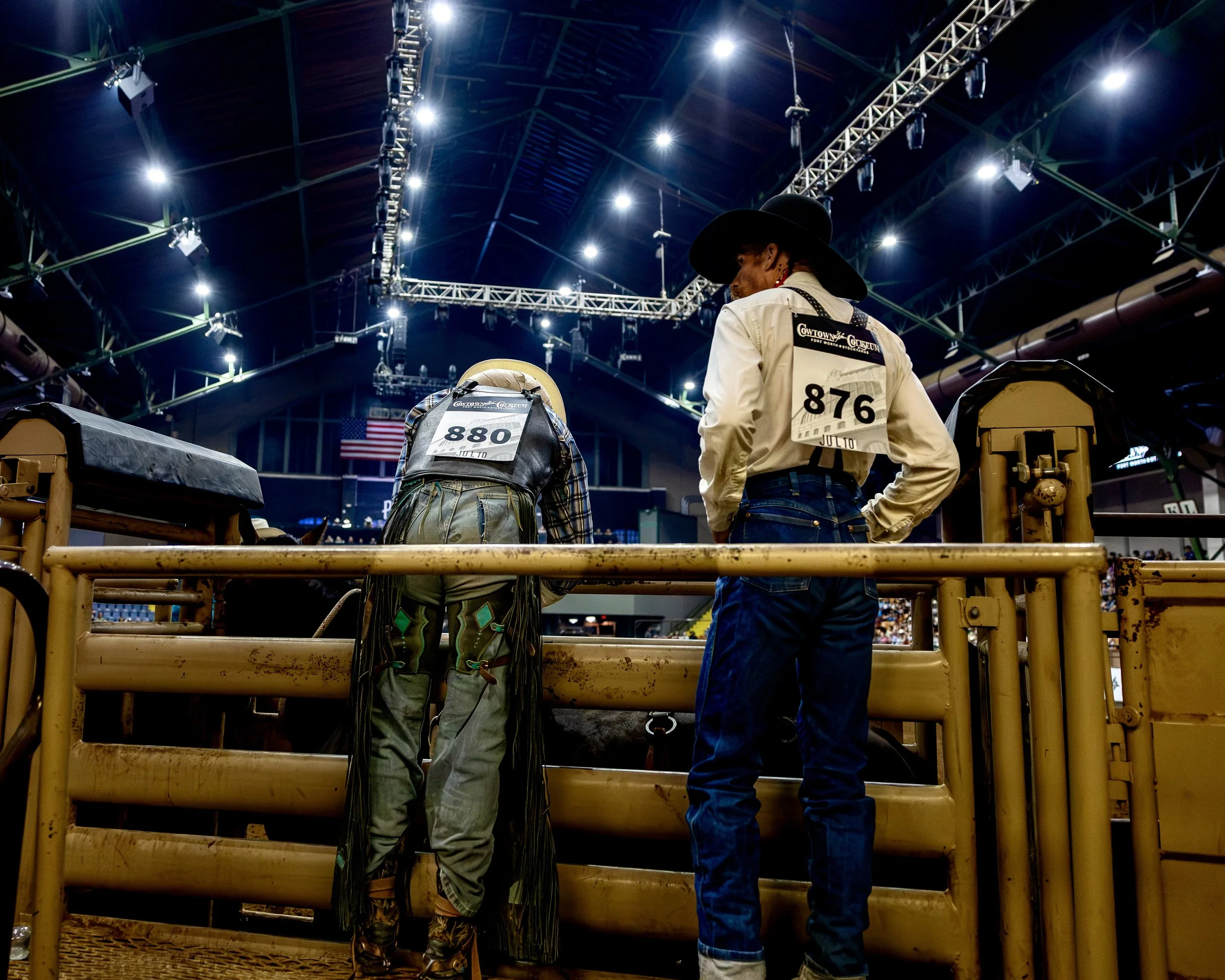 Two men in cowboy hats and western attire standing in a rodeo arena behind a yellow metal barrier, with bright overhead lights and a large indoor stadium structure in the background.