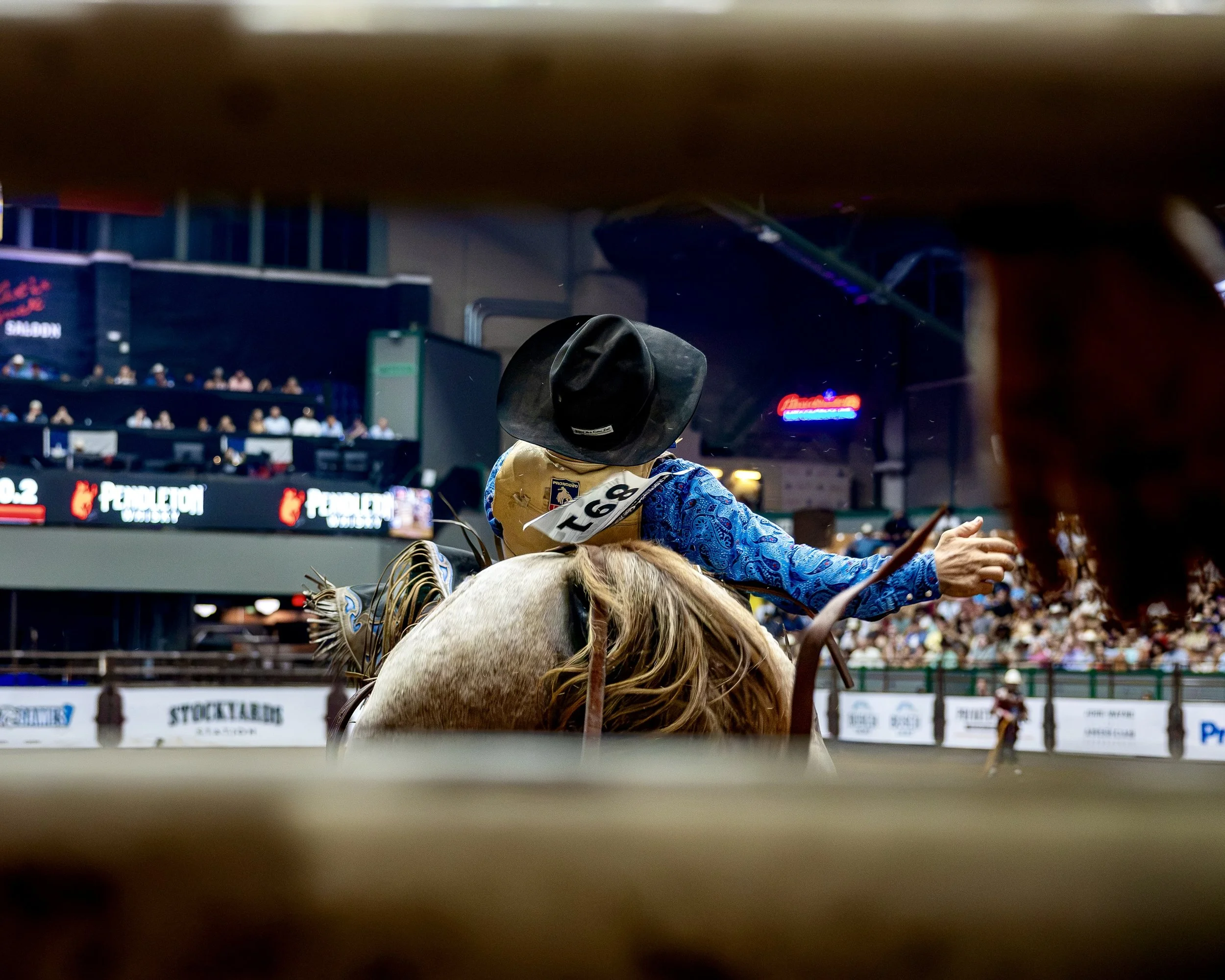 View from inside a rodeo arena showing a rider on a bucking horse, wearing a cowboy hat and colorful Western attire, with spectators in the background.