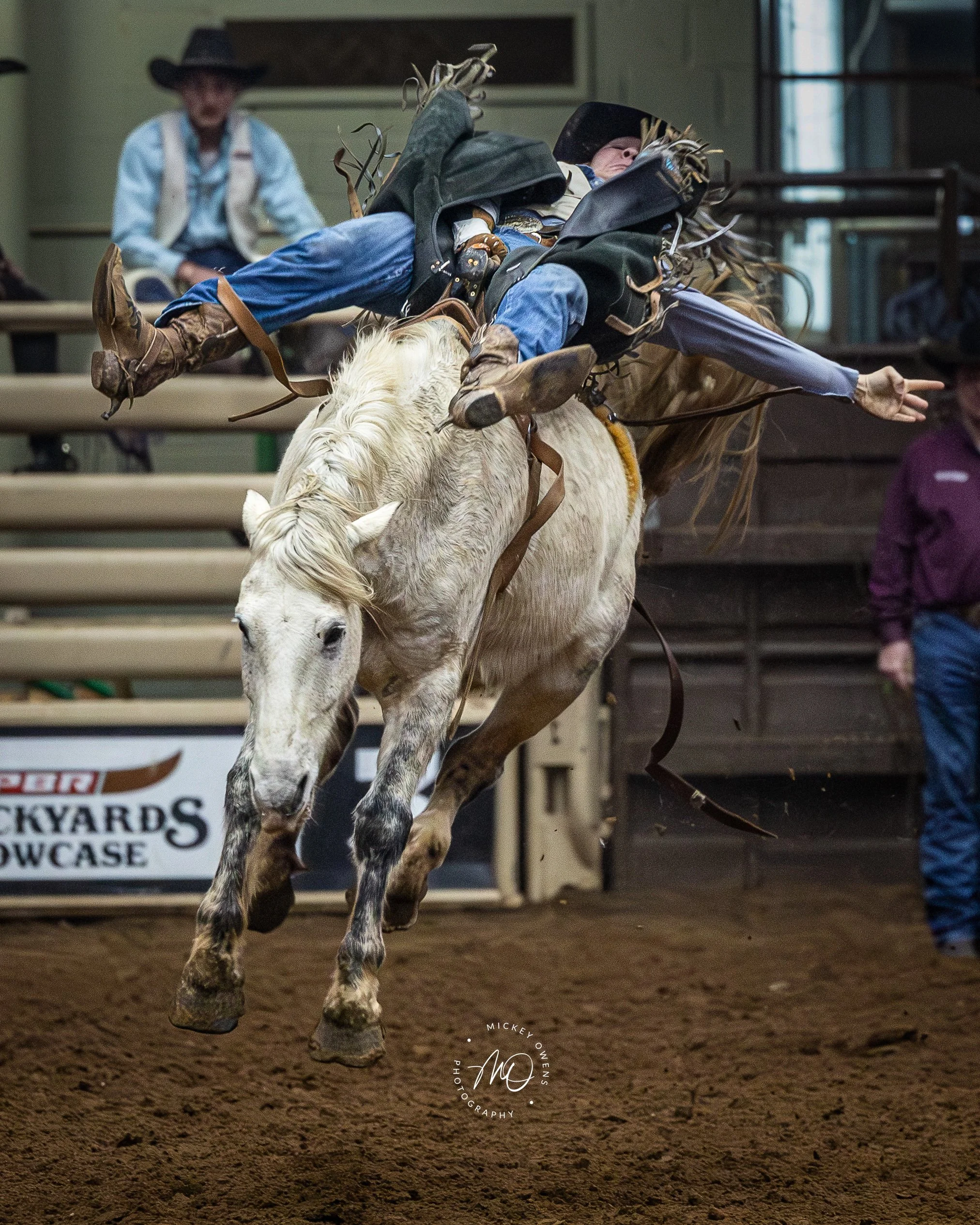 A rodeo cowboy riding a bucking horse on a dirt arena floor, with spectators in the background.