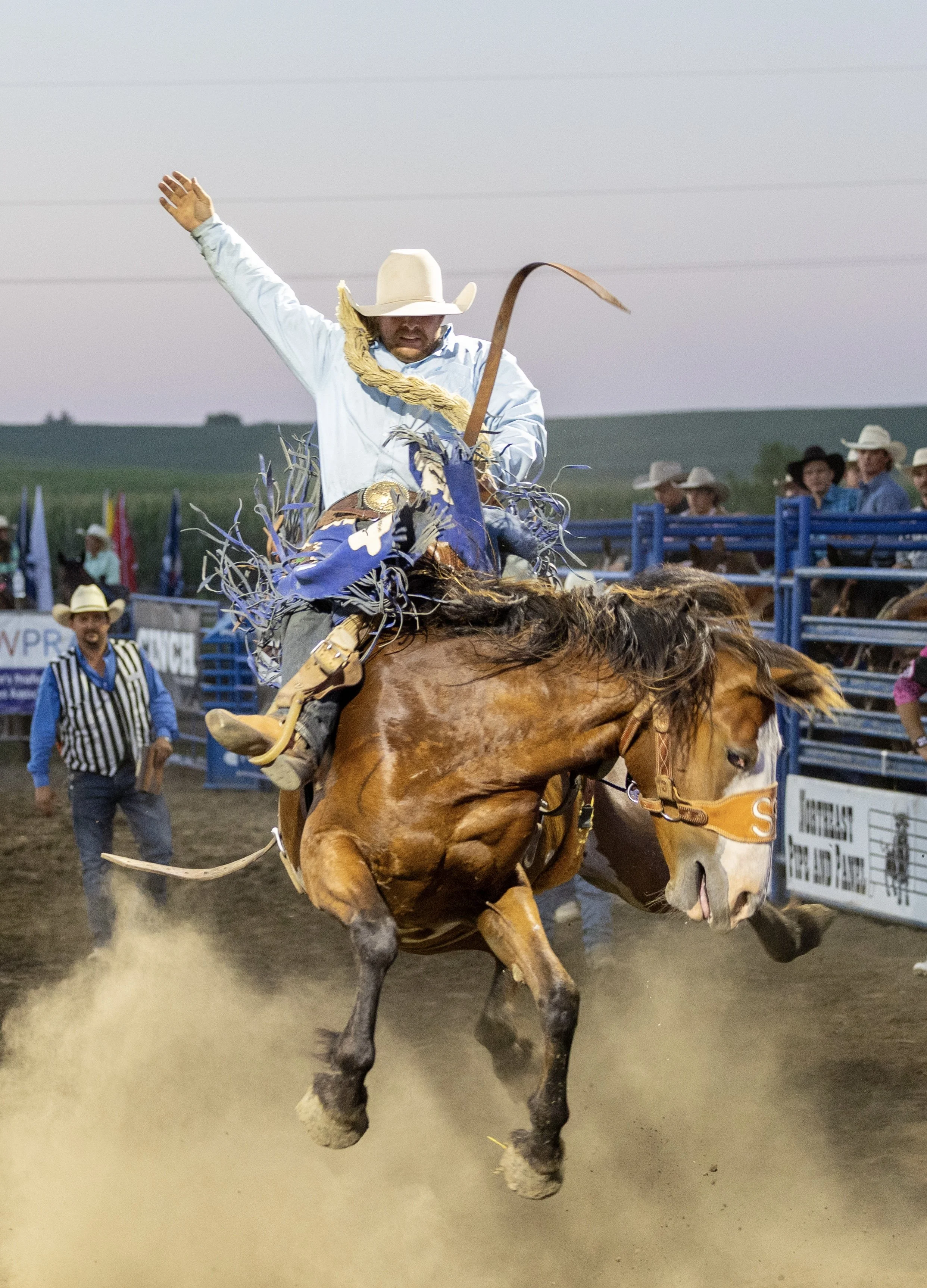A cowboy riding a bucking horse during a rodeo event, with spectators and officials watching in the background.
