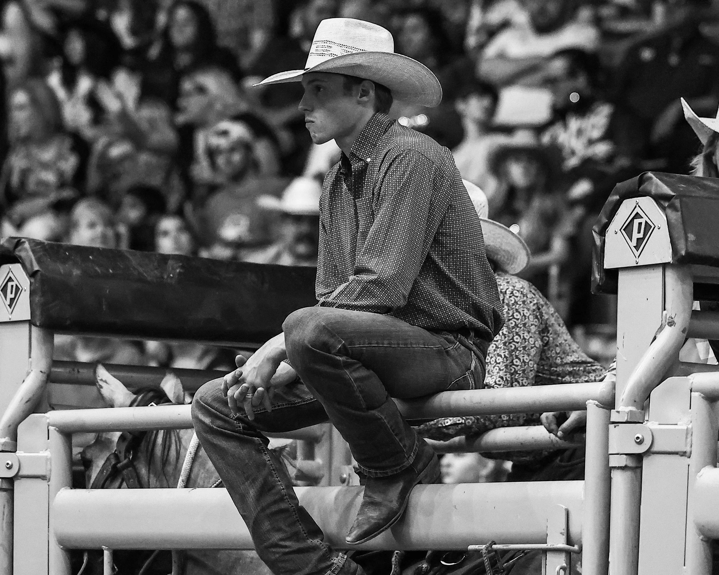 A young man wearing a cowboy hat and a checkered shirt sits on a bench, surrounded by a crowd of people, in an outdoor setting.