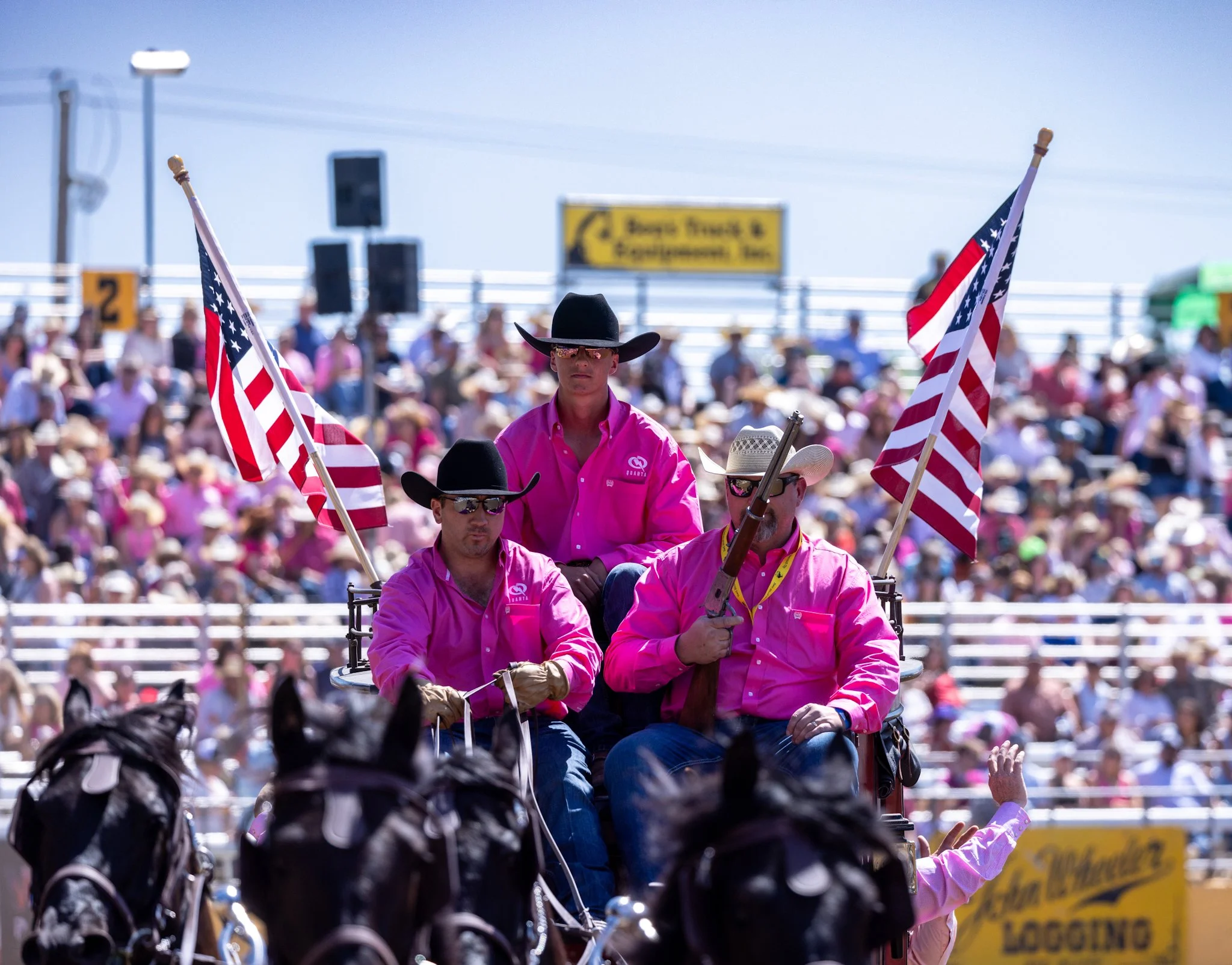 A parade scene featuring a group of people dressed in pink shirts, some wearing black cowboy hats, riding in a horse-drawn carriage with American flags. The crowd is visible in the background under a clear sky.