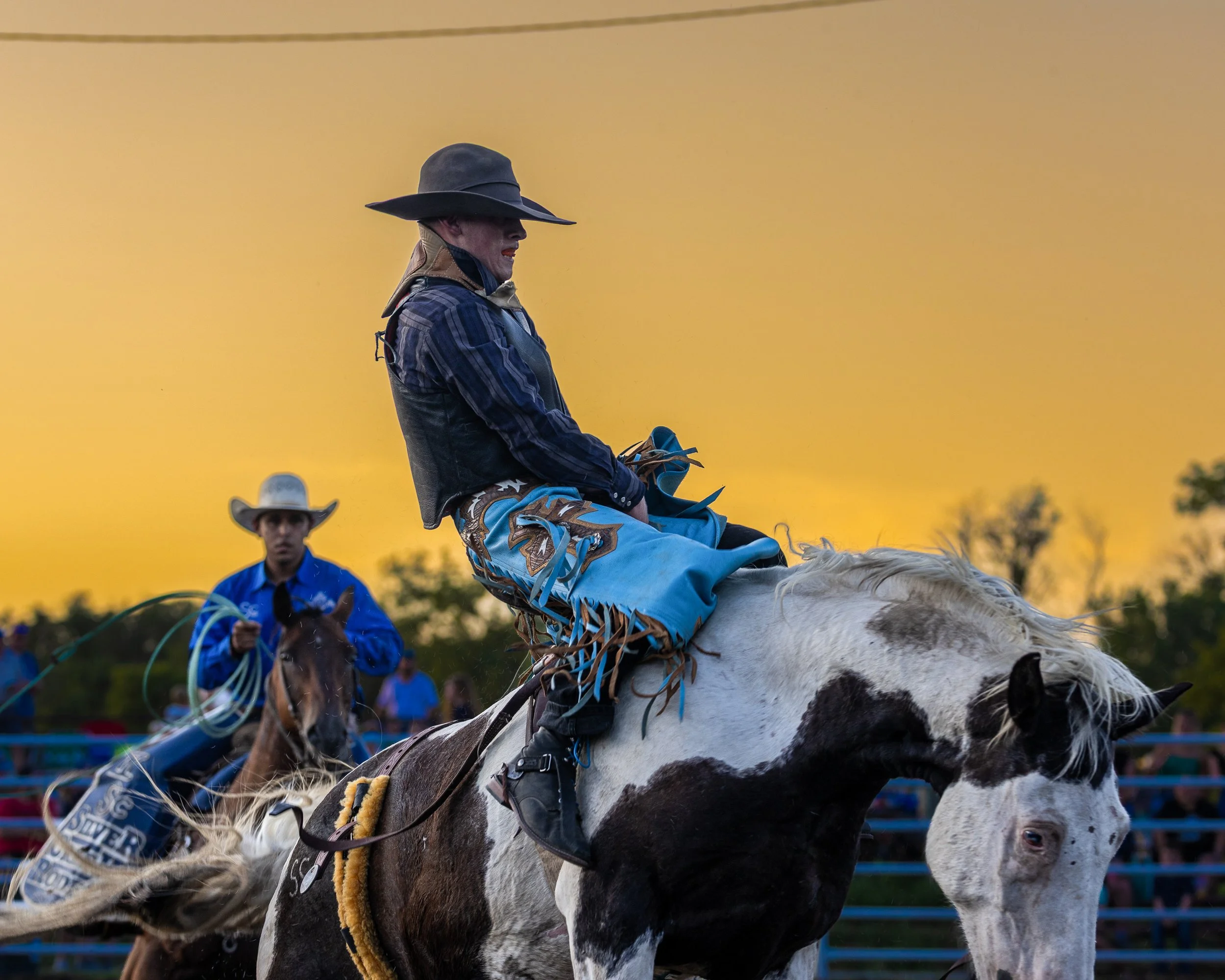 A man wearing a large black cowboy hat, riding a spotted horse during a rodeo event at sunset. There are other riders and spectators in the background.