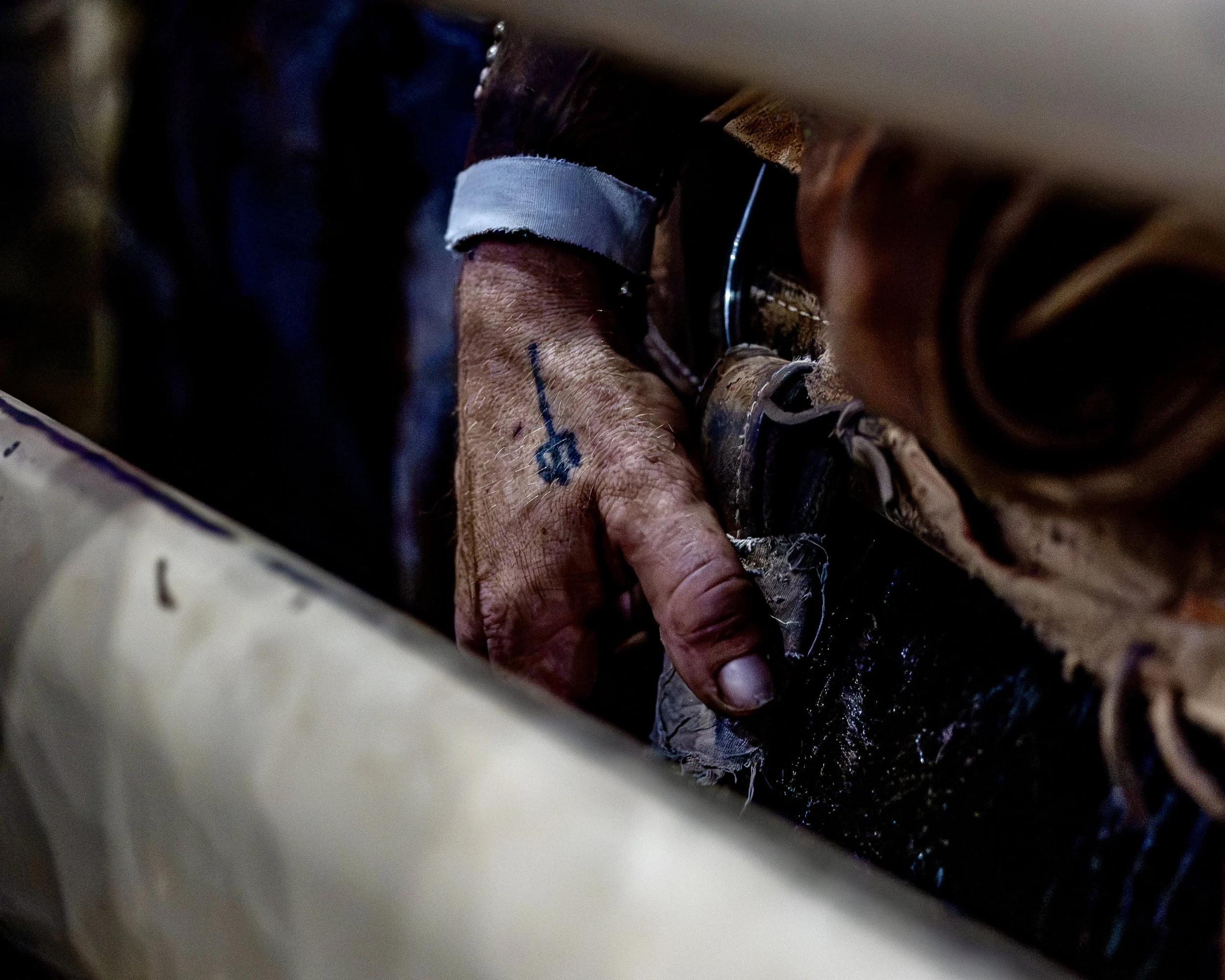 Close-up of an elderly person's hand holding a paintbrush, with a blue painted line on the finger. The background is dark and out of focus, with part of a striped fabric visible.