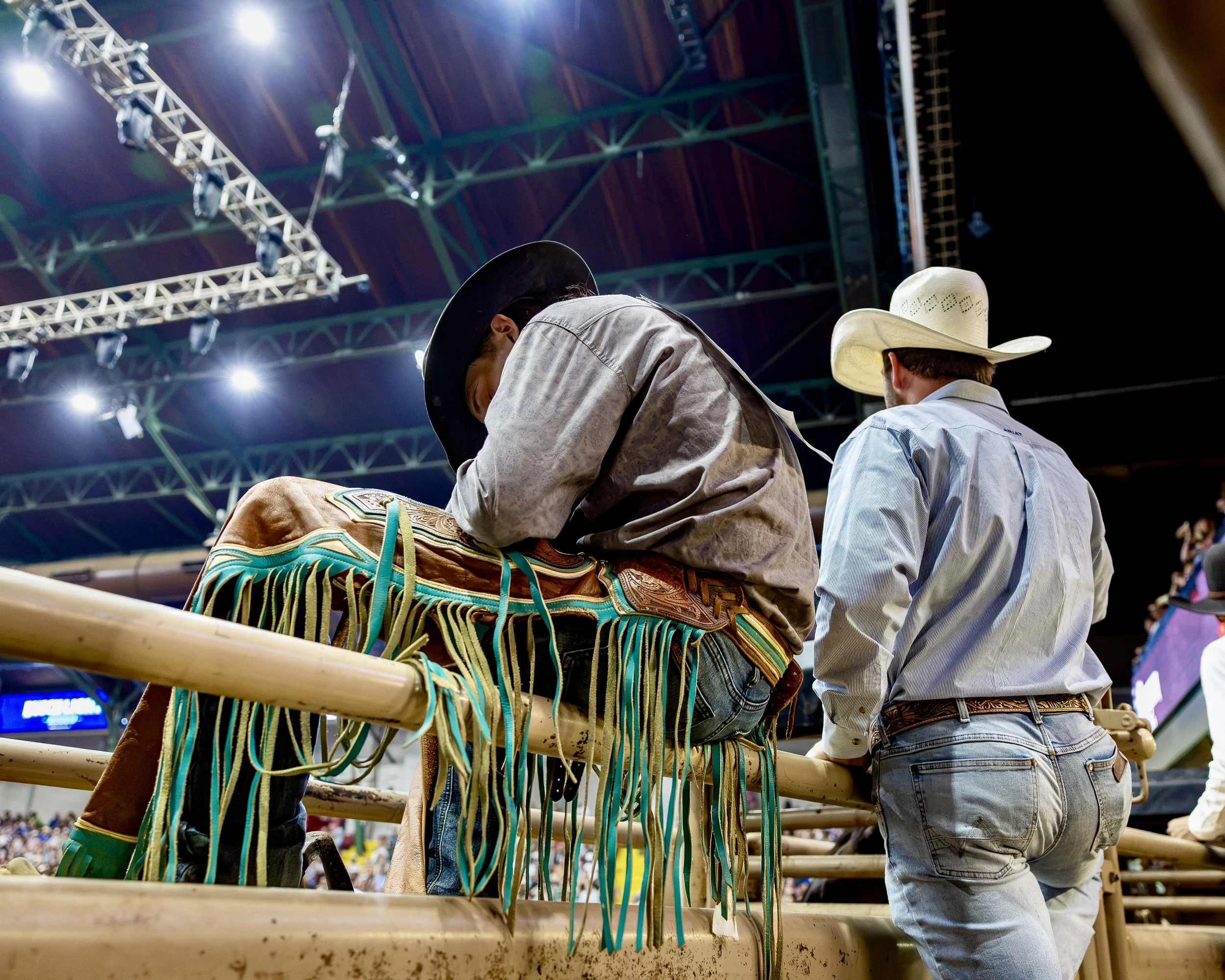 Two cowboys dressed in western attire are seen from behind, leaning over a fence or railing, amidst a large indoor arena with bright overhead lights and a high wooden ceiling, possibly during a rodeo event.