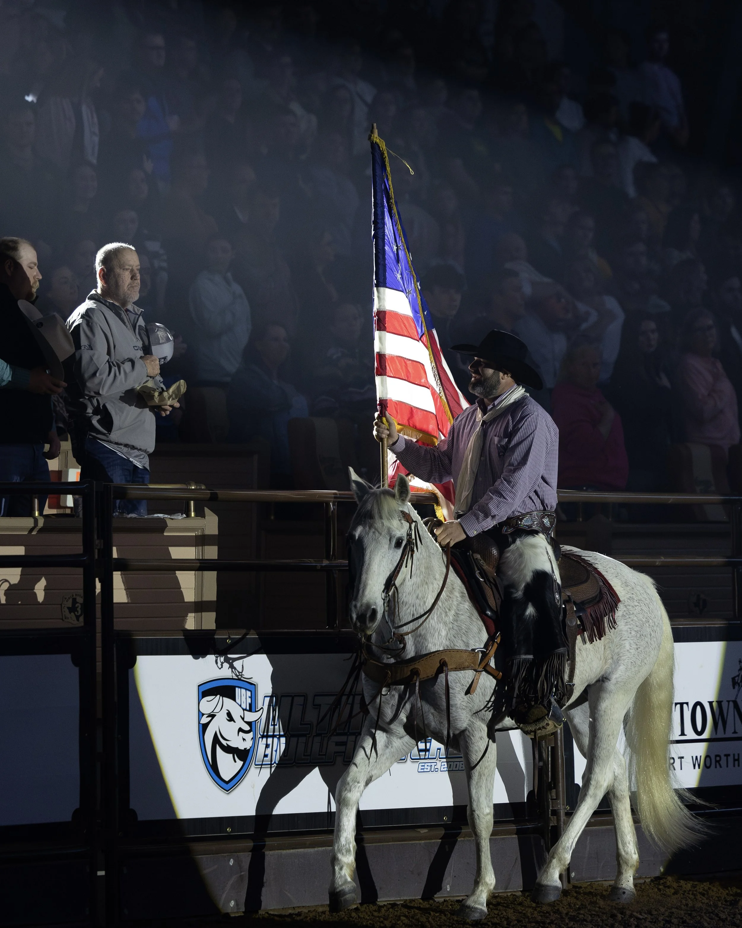 A man in western attire riding a white horse holding an American flag during a nighttime event with an audience in the background.