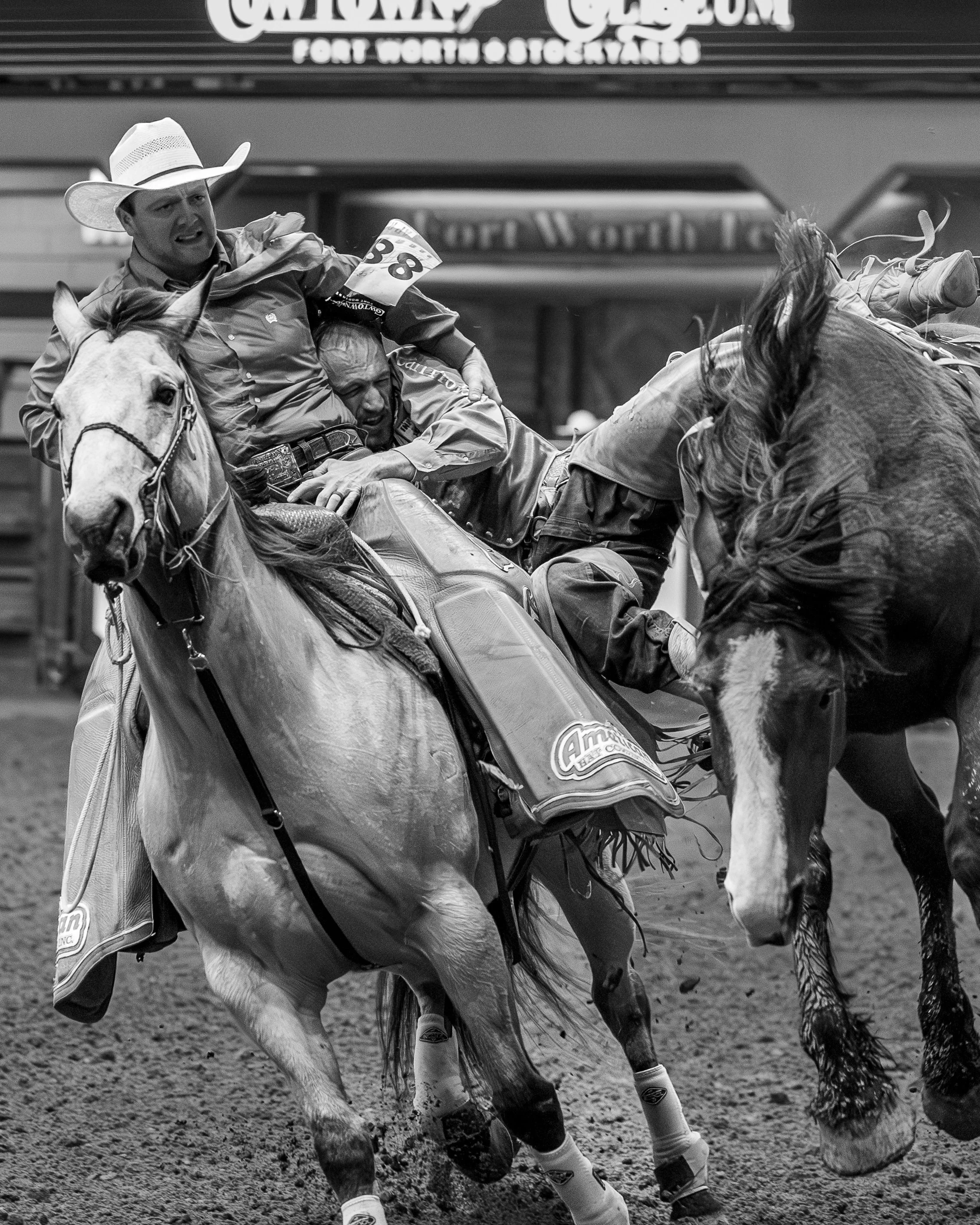 Two men competing in a rodeo event, riding horses and holding onto ropes, with one man wearing a cowboy hat and the other holding onto the horse. The scene appears intense as they are engaged in a rodeo event.