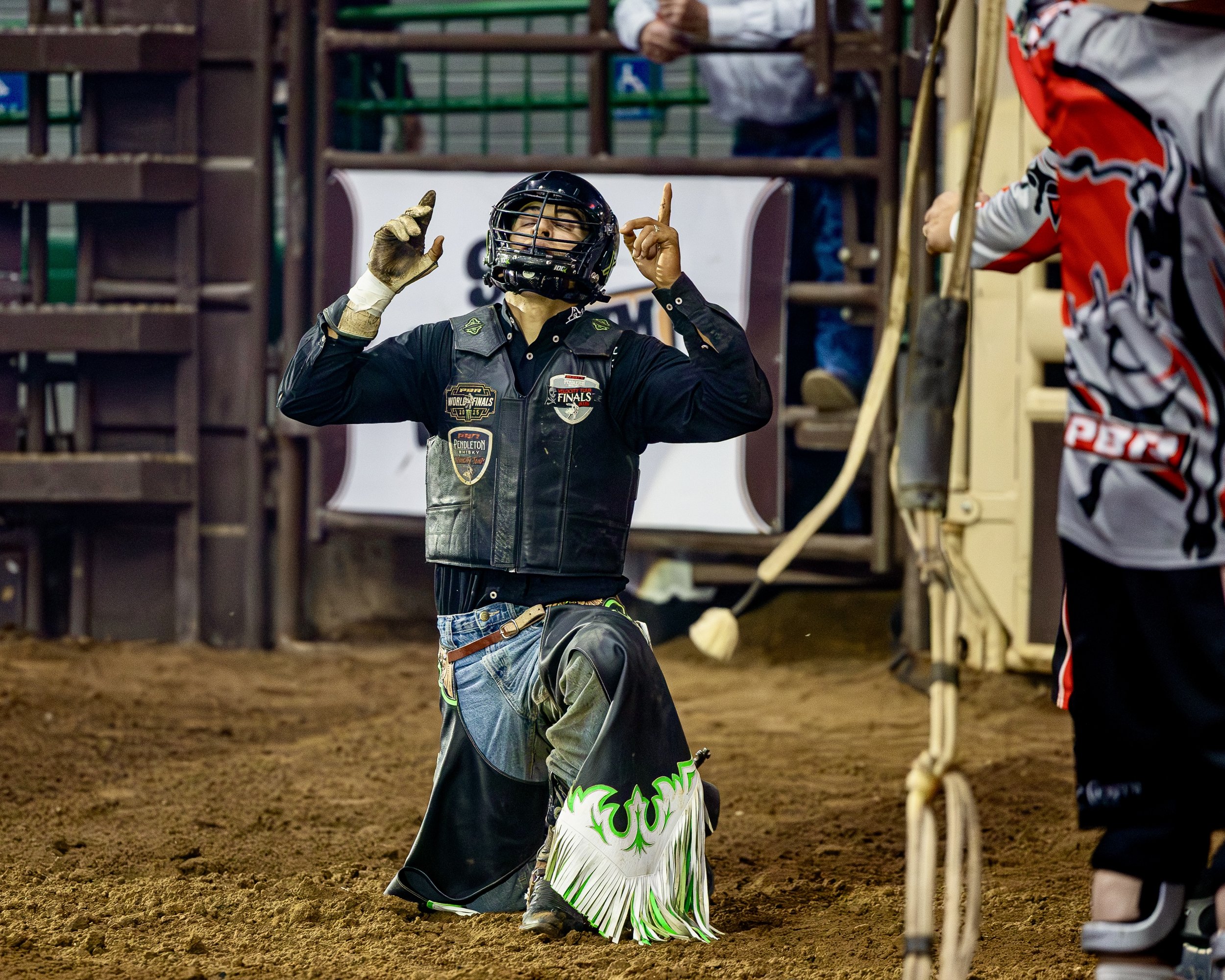 A rodeo clown wearing a black helmet and a leather vest with patches is kneeling on the ground and making a gesture with his hands. The background shows a gate, part of a bull riding arena, and other people, one wearing a red and black shirt.