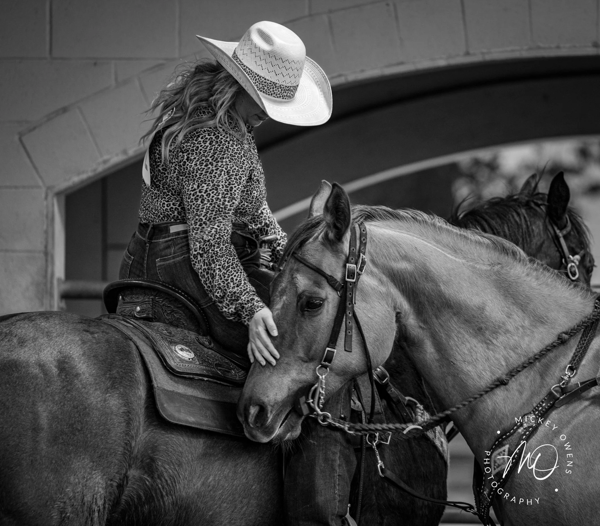 A woman wearing a wide-brimmed hat and a leopard-print shirt riding a horse indoors under a brick archway.