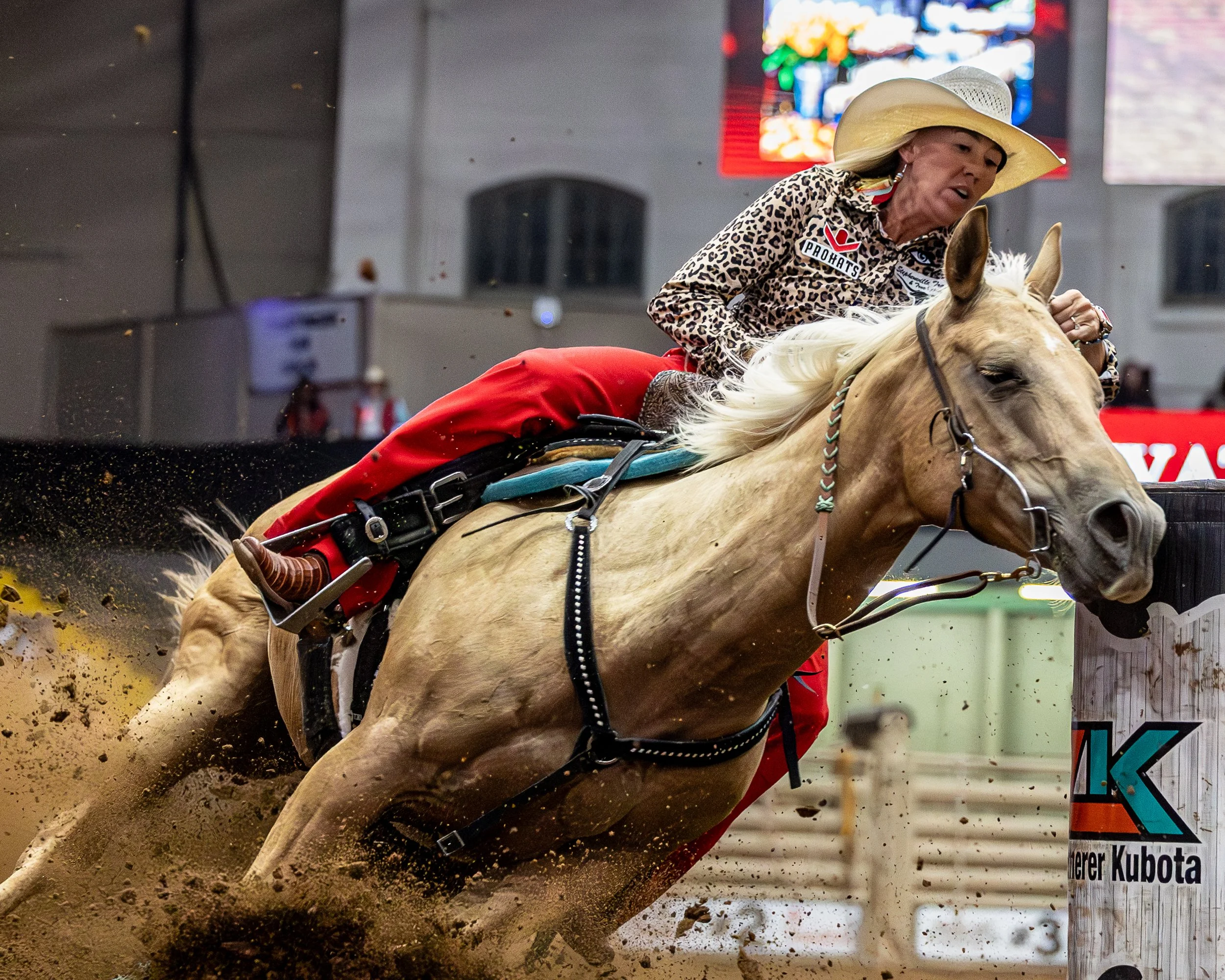A woman riding a light-colored horse during a rodeo, wearing a wide-brimmed hat, a leopard print shirt, and red pants, with a blurred background of an indoor arena and a sign.