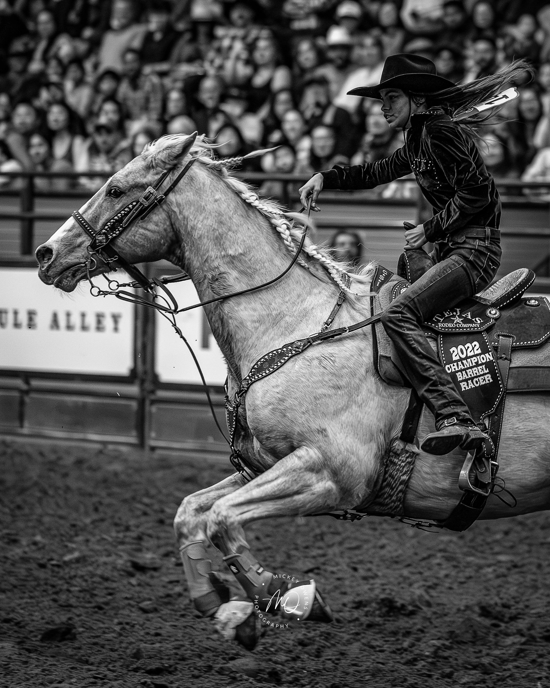 A female rodeo cowboy wearing a black hat and black western shirt rides a bucking horse during a rodeo event with a crowd in the background.