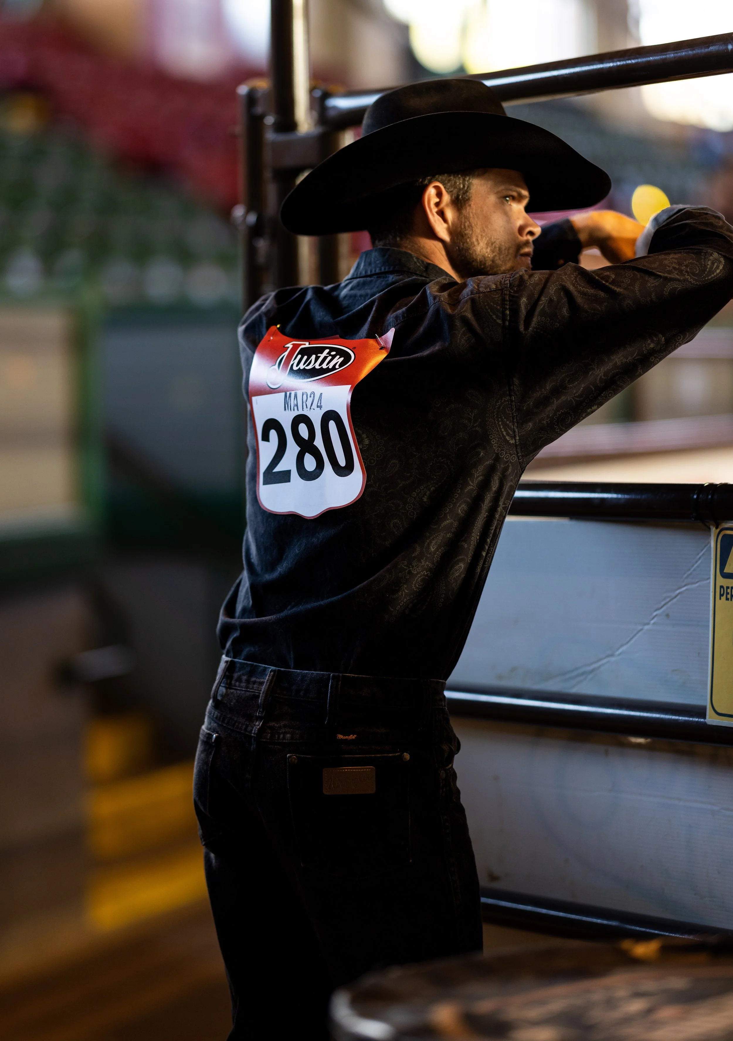 Man wearing a cowboy hat and black shirt, leaning on a metal railing in an indoor setting with a large white object and yellow caution sign nearby.