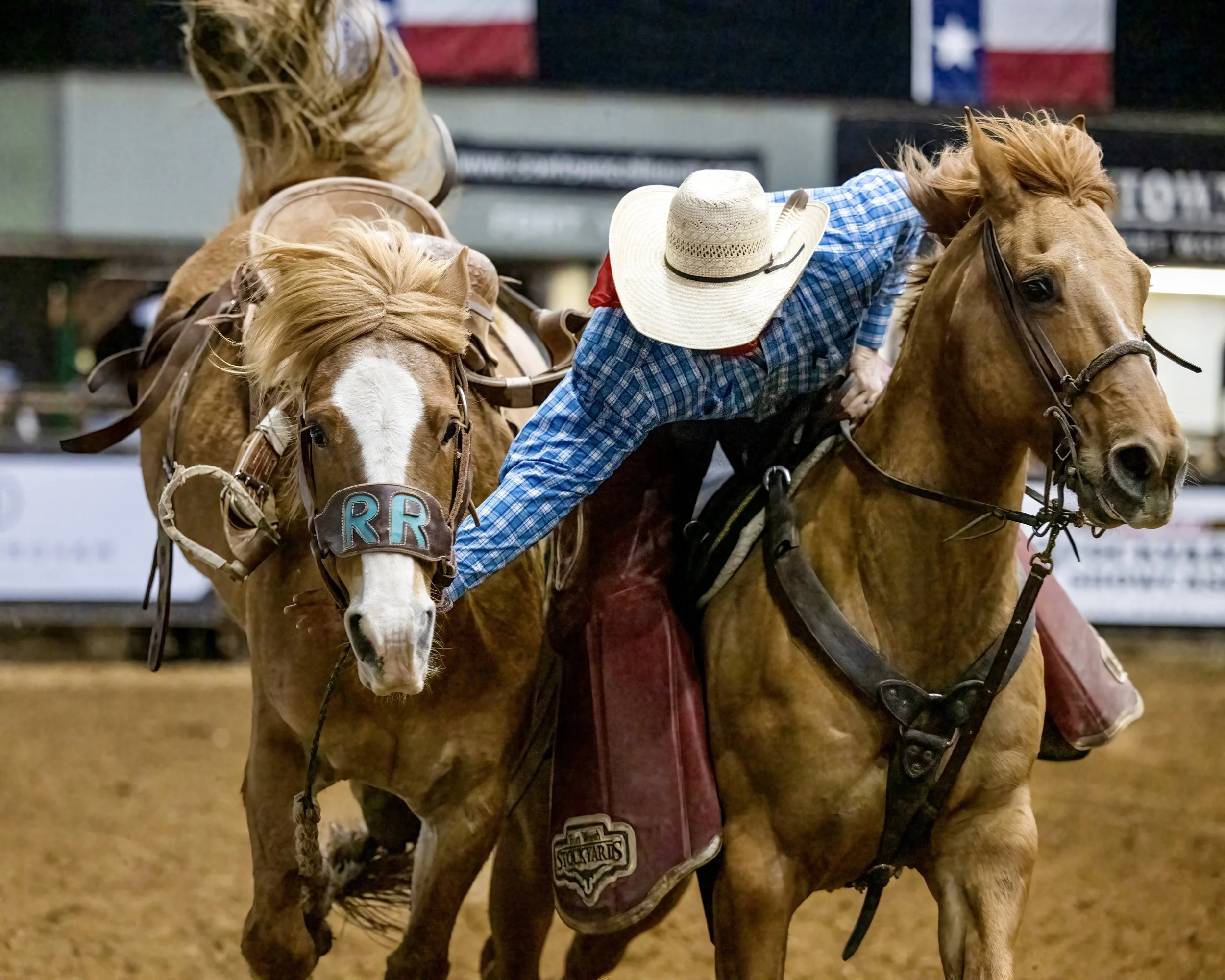Rodeo scene with two cowboys on horseback, one wearing a wide-brimmed straw hat and the other a cowboy hat, inside an arena with a blurred flag in the background.