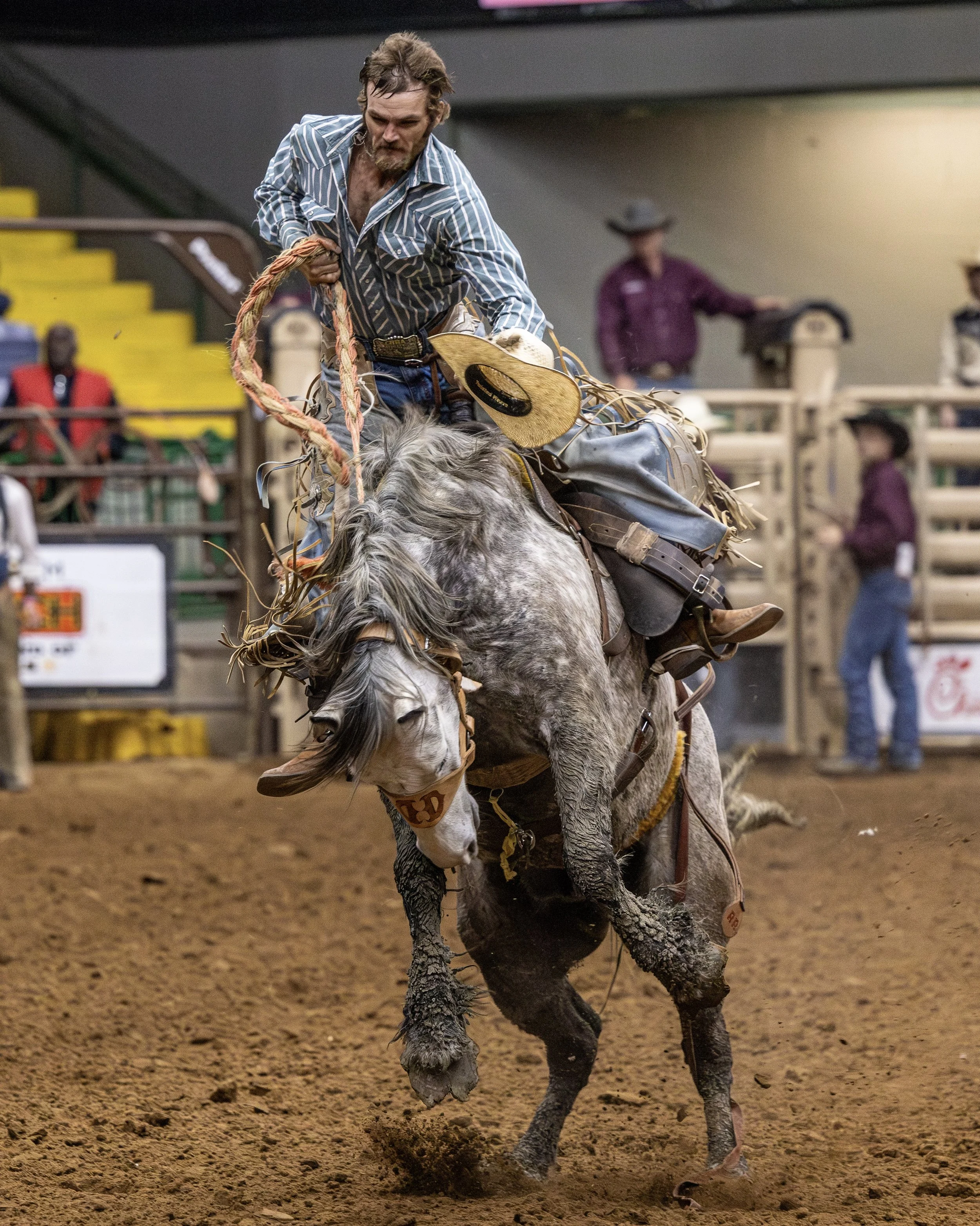 A cowboy riding a bucking horse during a rodeo event, with a dirt arena, spectators, and officials in the background.
