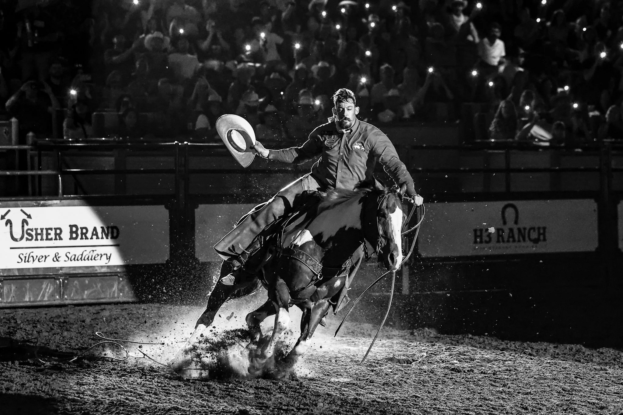 A cowboy riding a horse through water during a rodeo event, with a crowd of spectators in the background holding up lights or phones.