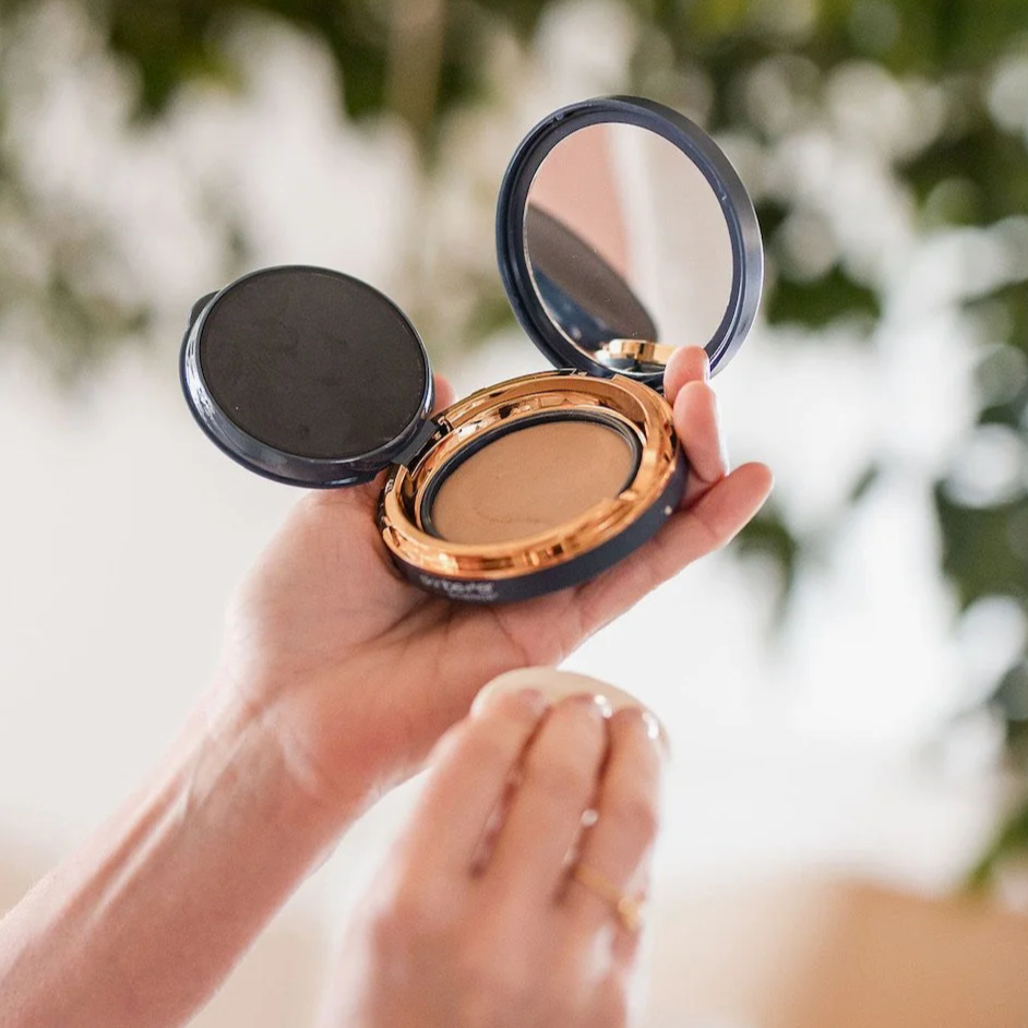 A hand holding an open compact powder with a mirror, against a blurred background of green and white.