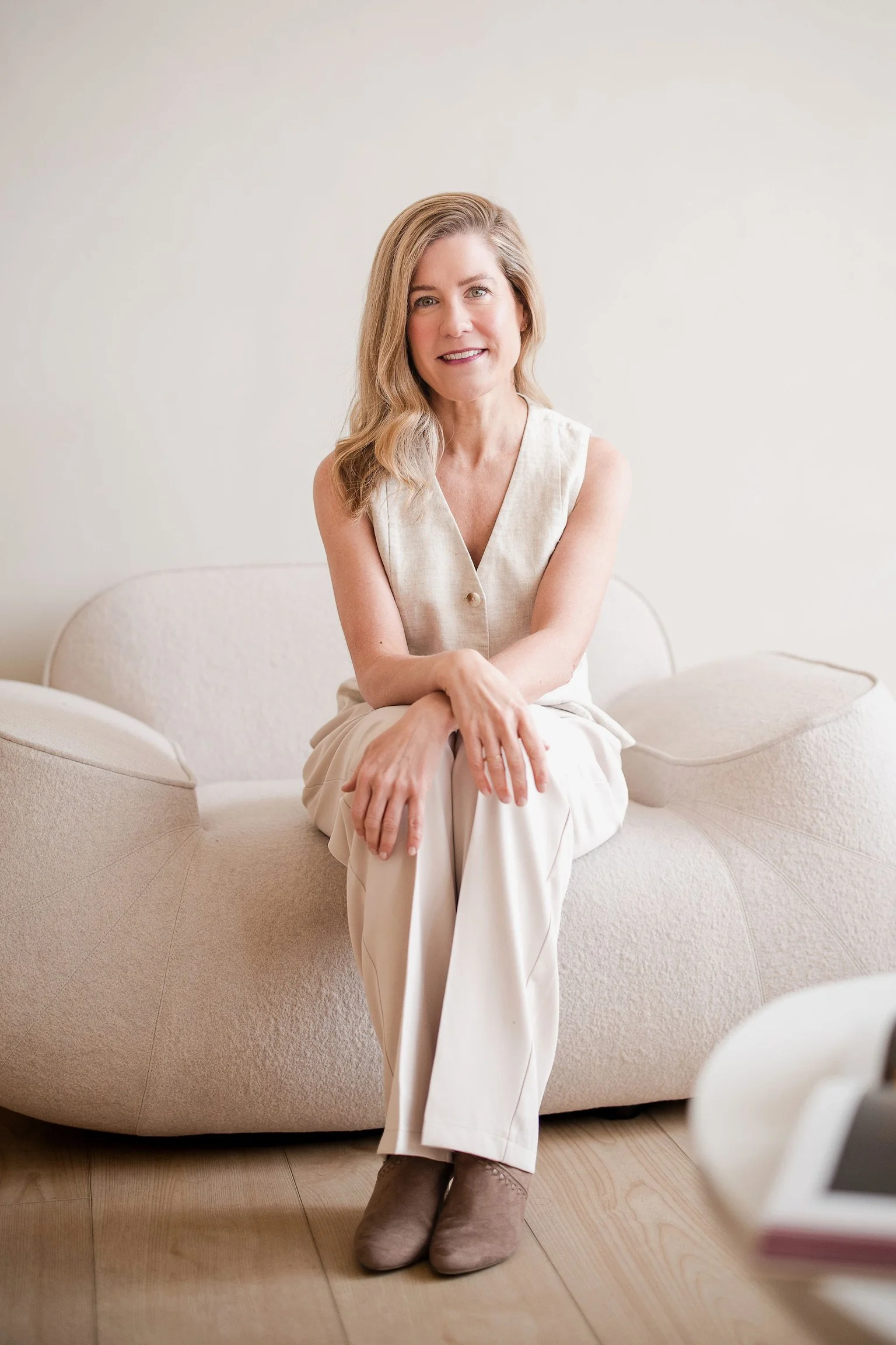 A woman with blonde hair, wearing a light sleeveless top and beige pants, sitting on a beige curved sofa in a minimalistic room with wood flooring.
