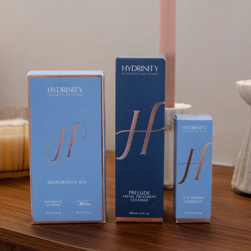 Three skincare product boxes from Hydrinity on a wooden surface. The left box is a Restorative Kit, the middle is a Prelude Facial Treatment Cleanser, and the right is an Eye Renew Complex.