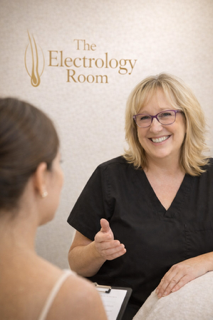 A woman with blonde hair and glasses smiling and talking to a patient in a clinical setting with a sign that says 'The Electrology Room' in the background.
