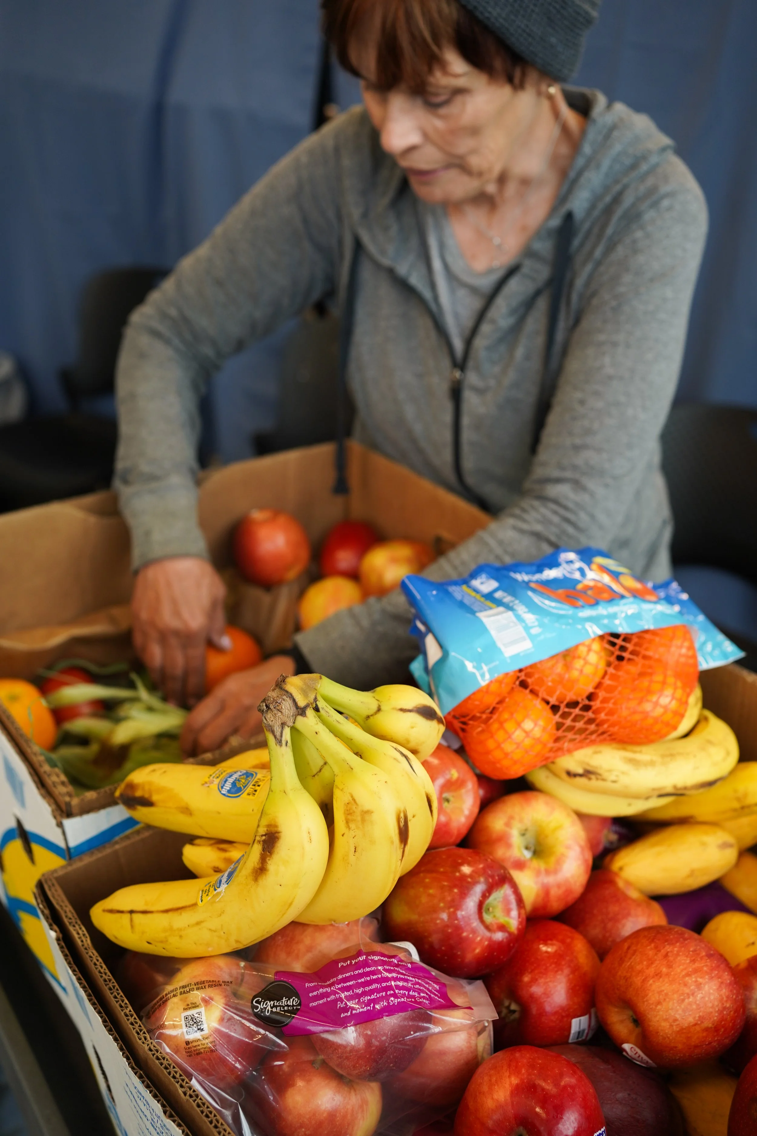 A woman wearing a gray hoodie and a beanie selecting apples and bananas from a box of assorted fresh fruits including red apples, bananas, and oranges at the food bank