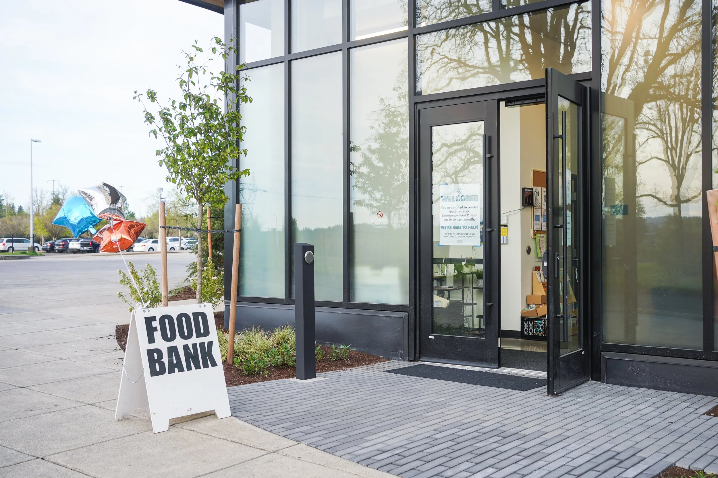Exterior of the Wilsonville Community Sharing facility with glass walls and an open door, sign on door, and a sidewalk. Outside, there is a sign that reads "FOOD BANK", a couple of balloons, and a small tree.