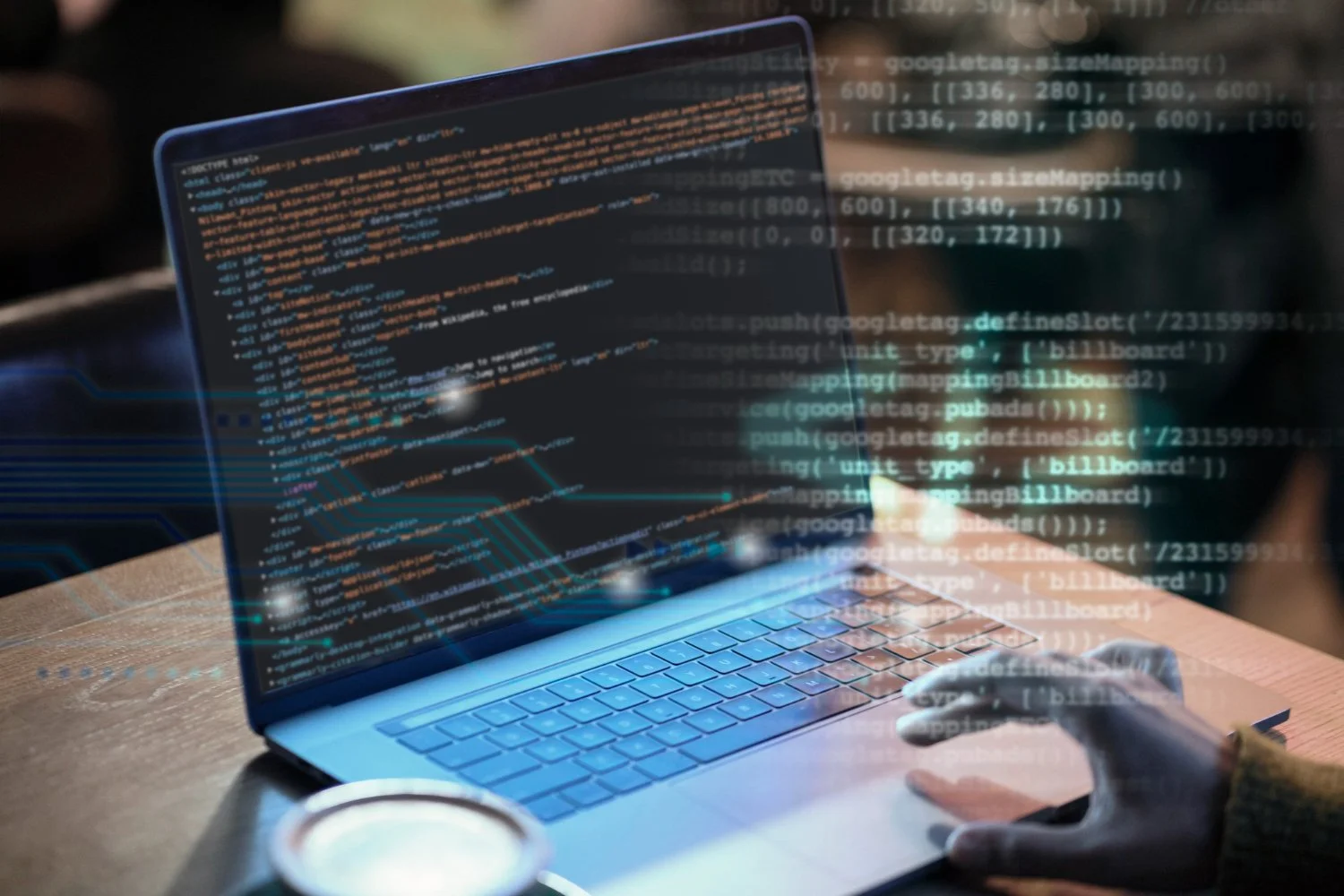 A person using a laptop with visible code on the screen, with a holographic display of programming code overlayed in front of the laptop, sitting at a wooden desk.