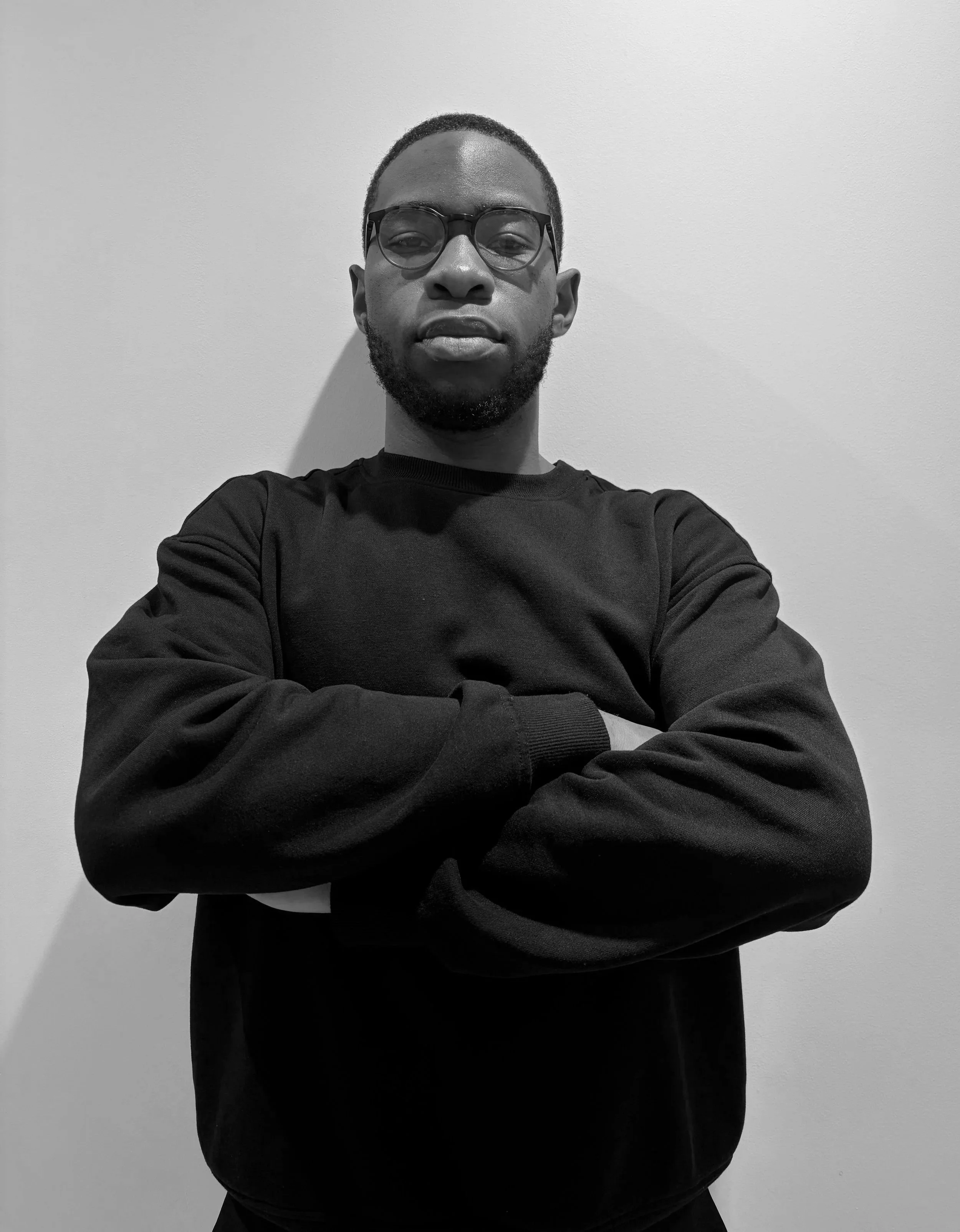 A black and white portrait of a man wearing glasses and a black long-sleeve shirt, standing against a plain wall with arms crossed.