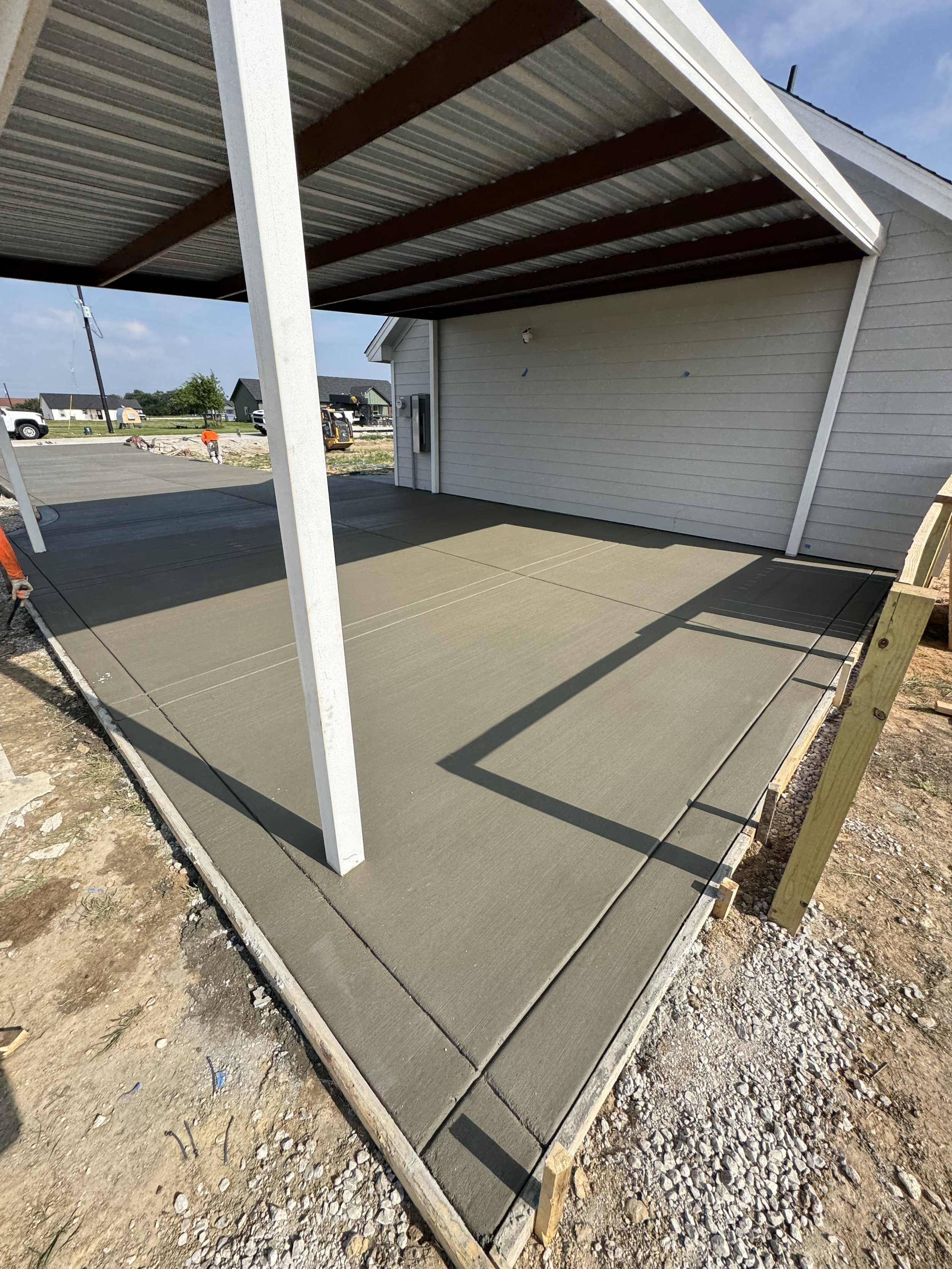The photo shows a newly poured concrete slab foundation for a building, with wooden forms and rebar visible along the edges. The site is outdoors with some construction workers and equipment in the background, and a partially constructed building structure overhead.
