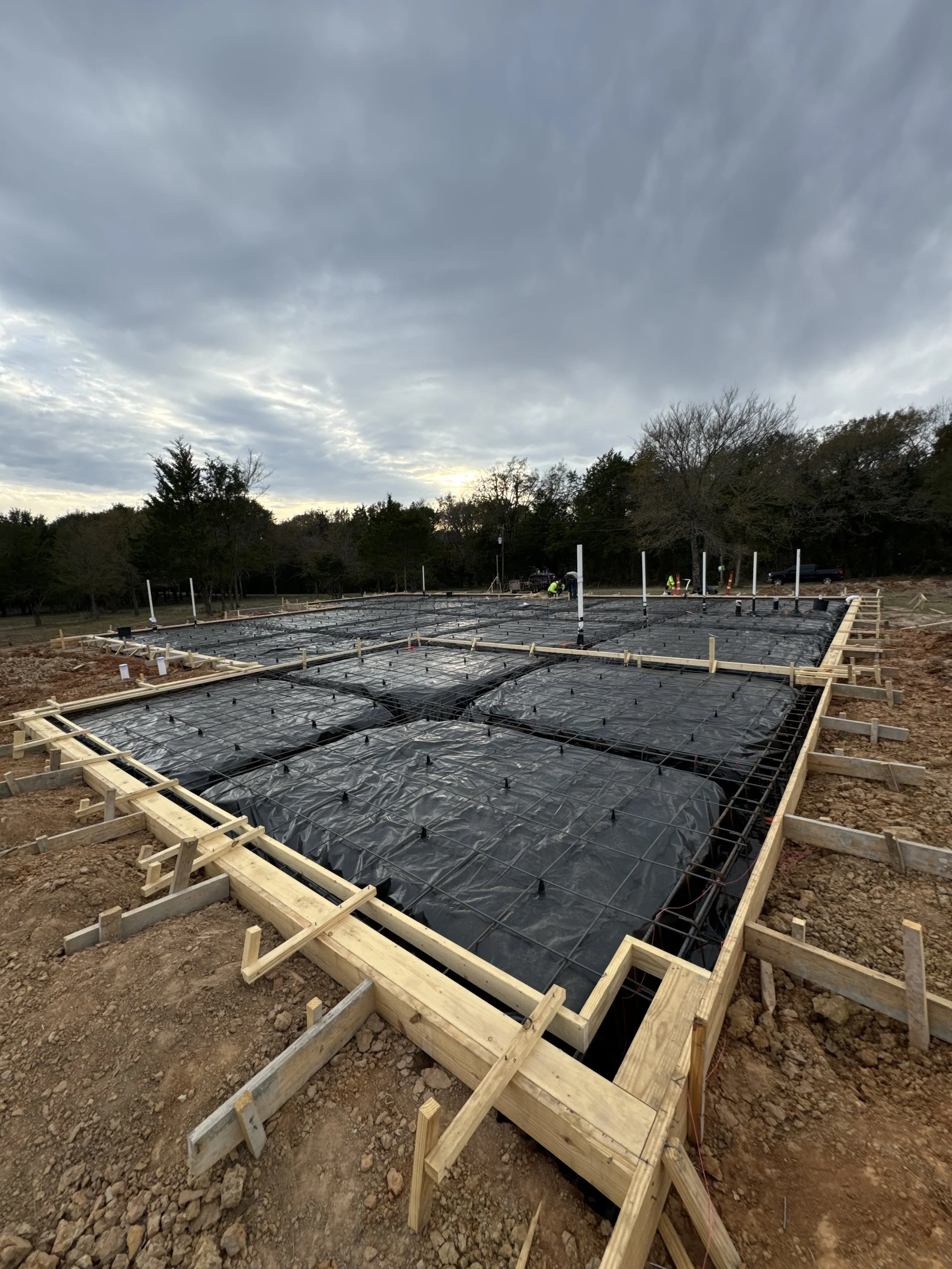 Construction site showing foundation work with black plastic sheeting, wooden framing, and steel reinforcement bars, set against a cloudy sky and trees in the background.