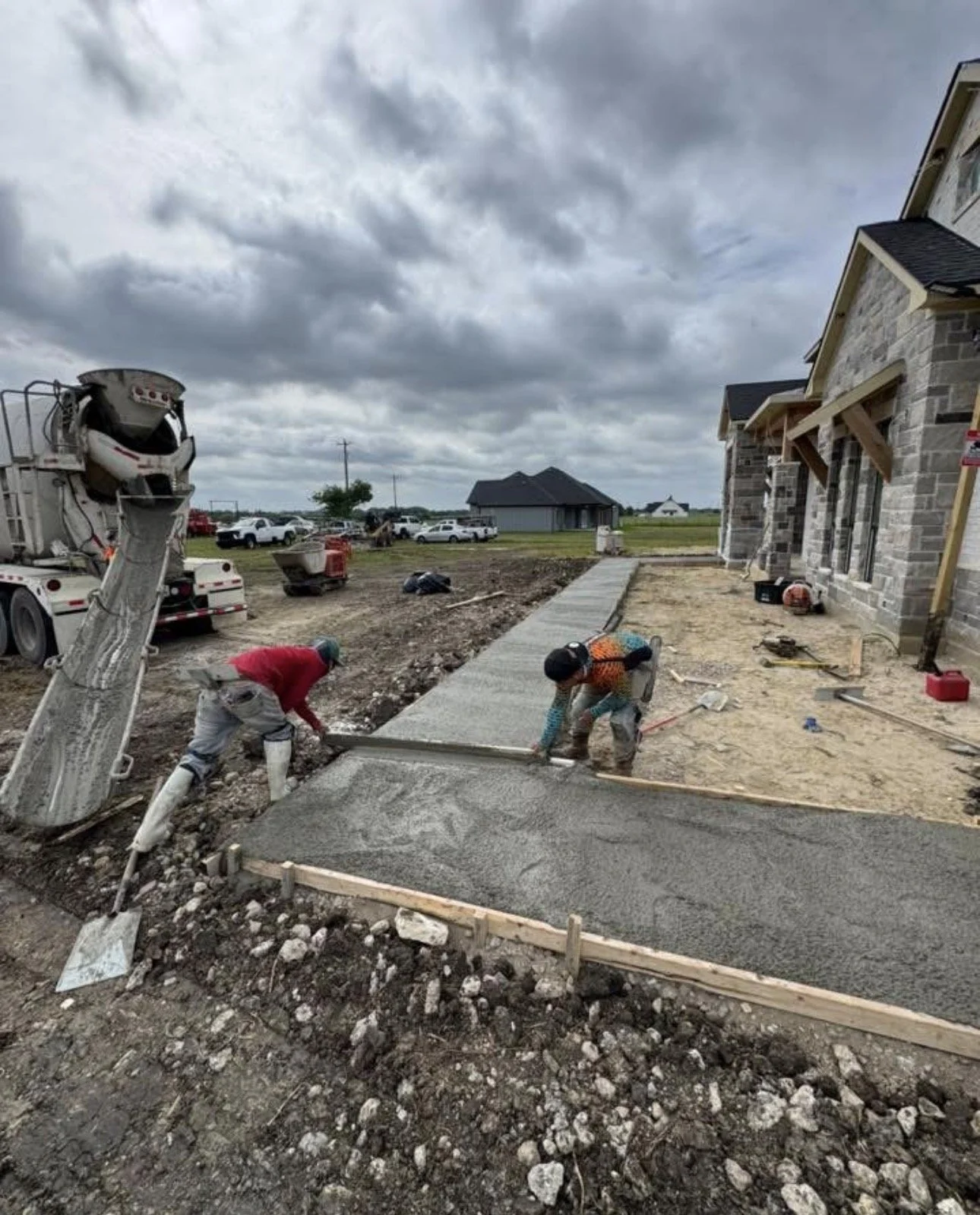 Construction workers pouring and smoothing concrete for a sidewalk in front of a house under construction, with clouds over a semi-rural area.