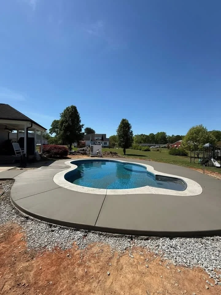 A backyard with a kidney-shaped swimming pool, surrounded by a concrete deck, with trees and houses in the background under a clear blue sky.