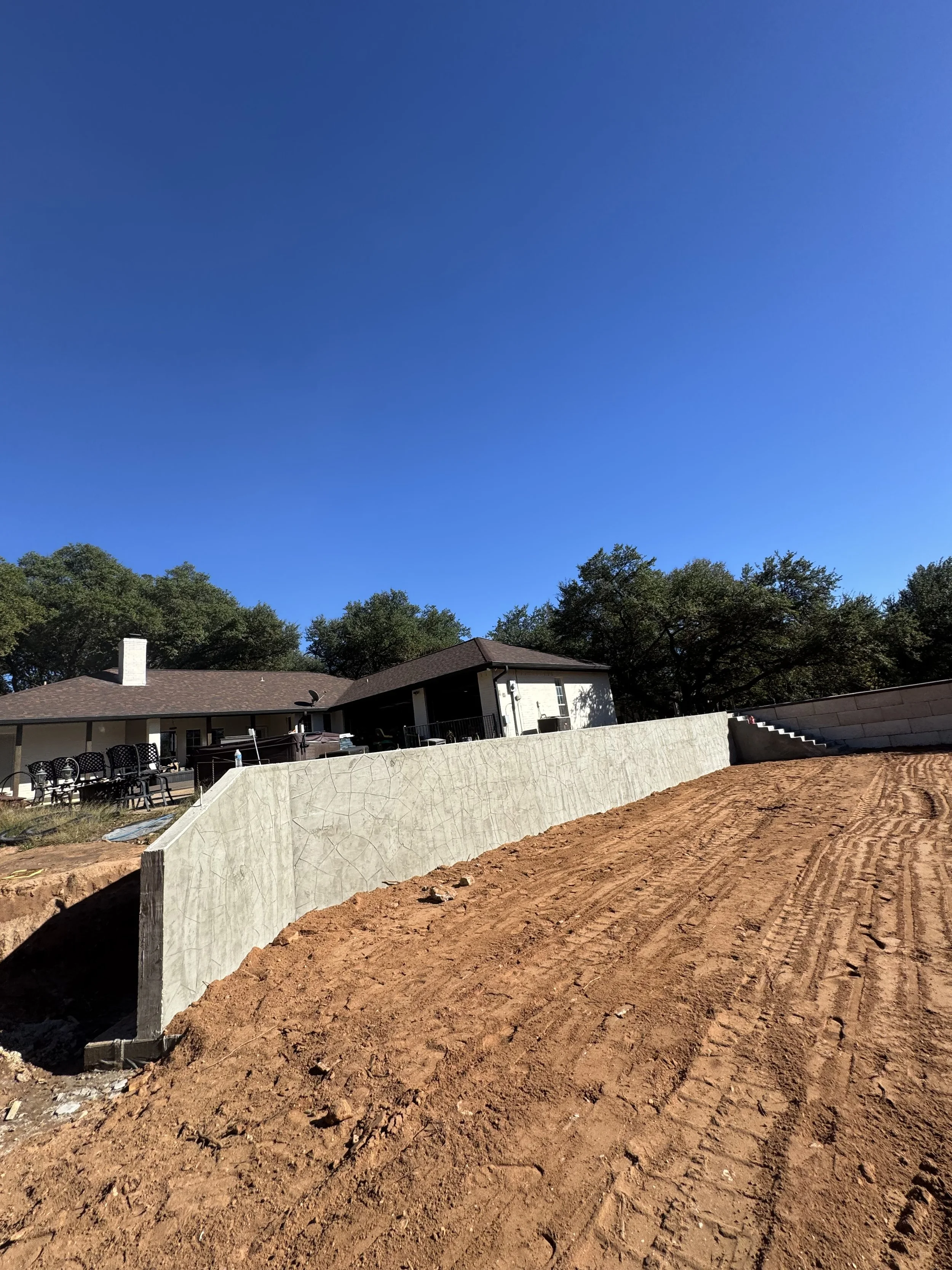 A backyard under construction with a concrete retaining wall, dirt ground, and a house with a brown roof and chimney, surrounded by trees and a clear blue sky.