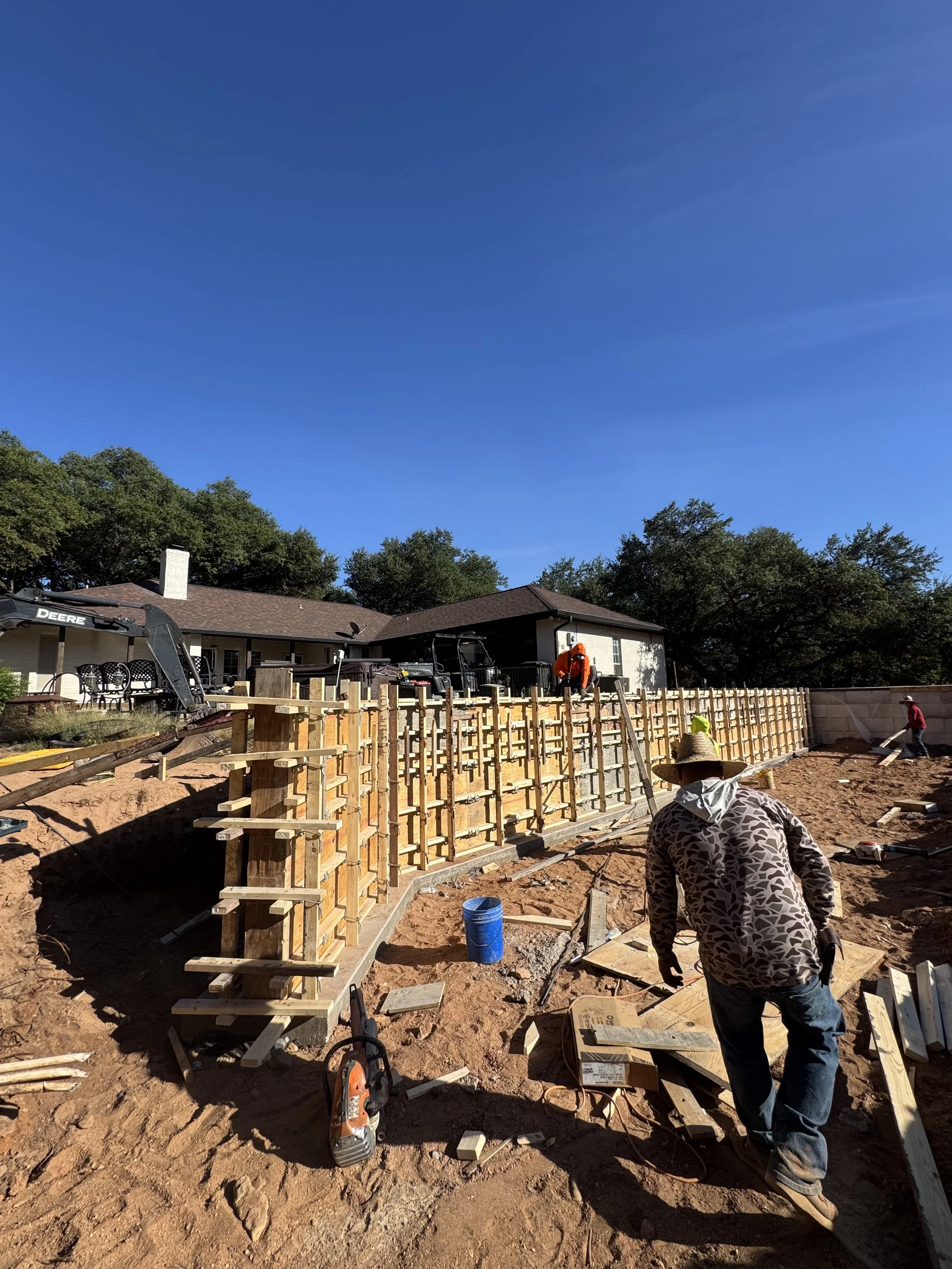 Construction workers building a concrete foundation wall in a backyard on a sunny day, with a house and trees in the background.