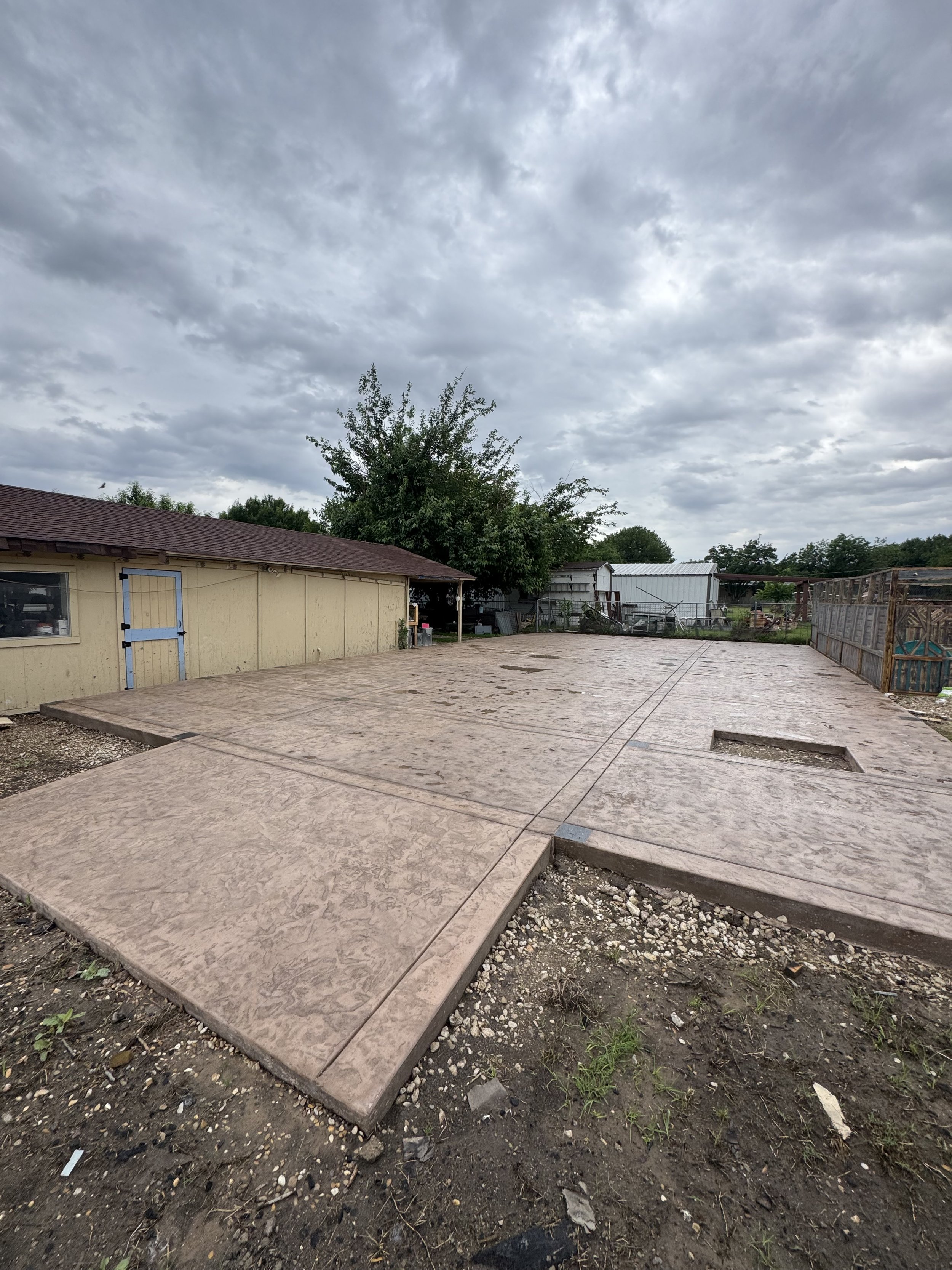 Construction site with freshly poured concrete slabs, a small building with a blue door, a tree, and a cloudy sky.