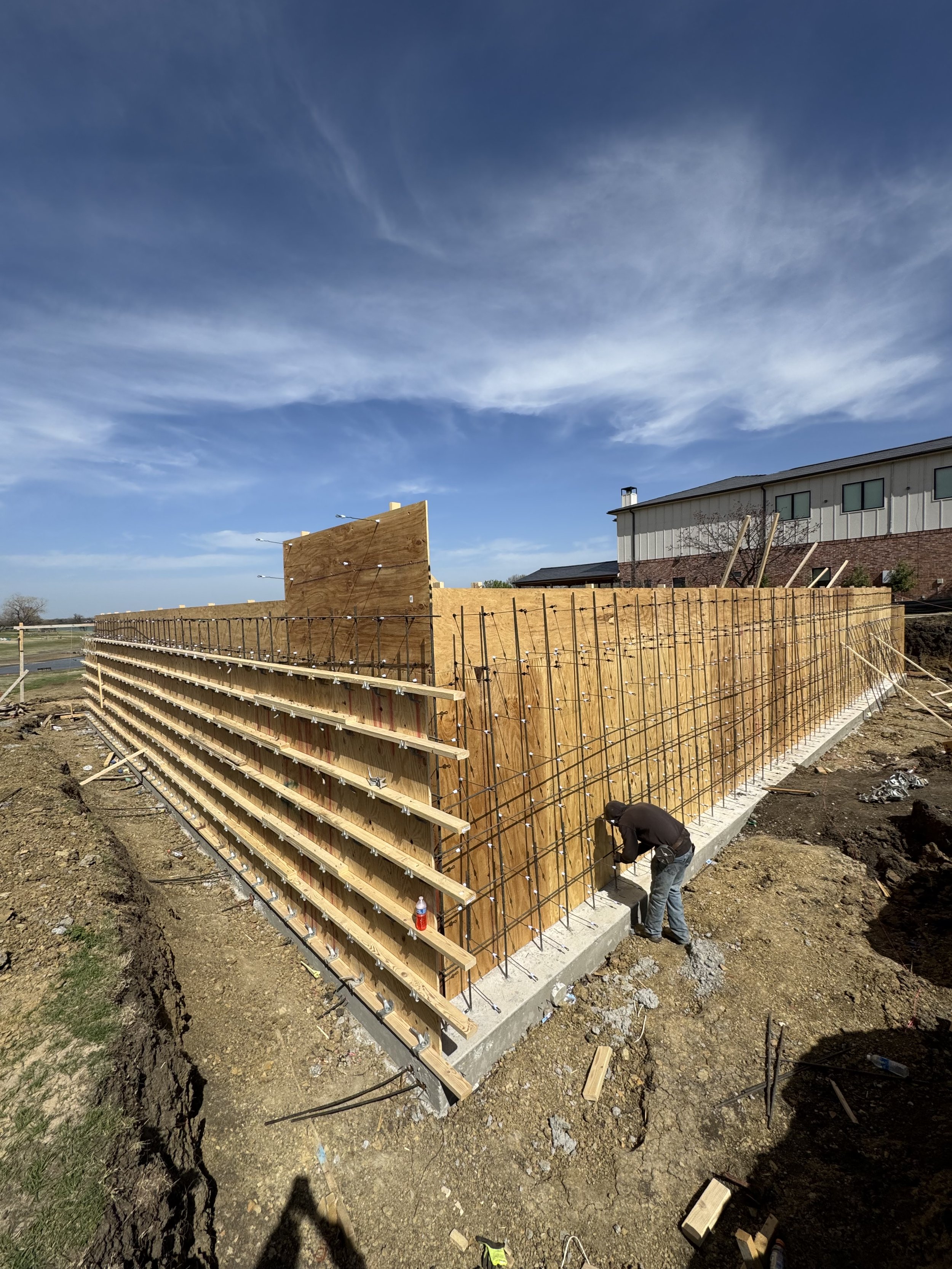 Construction site with wooden framing and rebar, a worker adjusting the concrete foundation, and a building with large windows in the background.