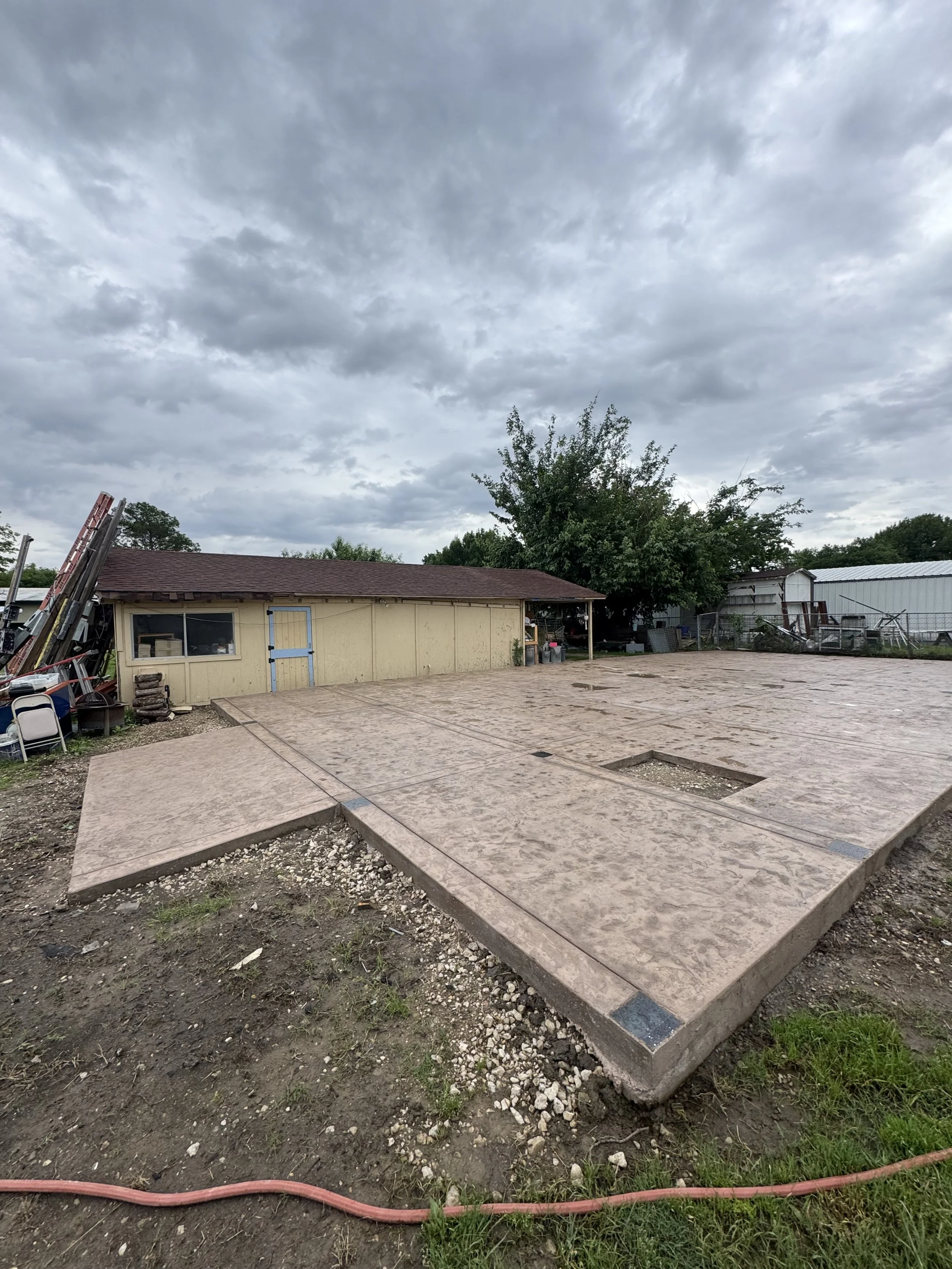 A small yellow building with a red roof, in a backyard with trees and a metal shed. The ground is being prepared for concrete, with a leveled area and some gravel, and a red hose lies on the dirt in the foreground.