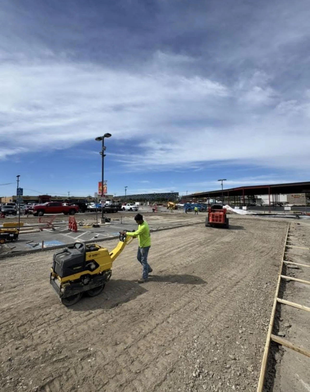 Construction site at a shopping plaza with workers, heavy machinery, and partially paved ground, under a cloudy sky.