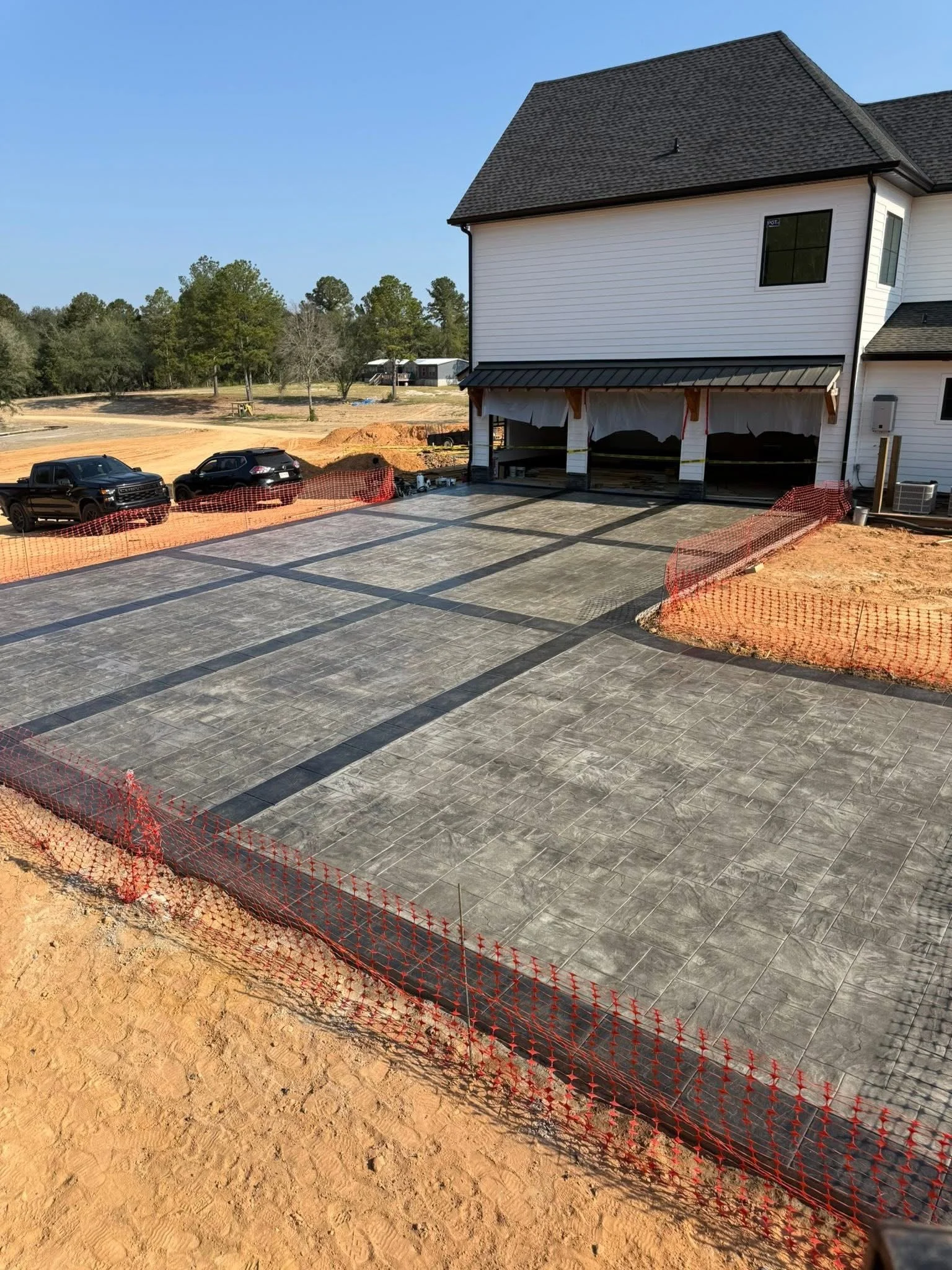A residential construction site with a newly paved driveway, partially surrounded by orange safety fencing, next to a white house with black roof shingles, under a clear blue sky.