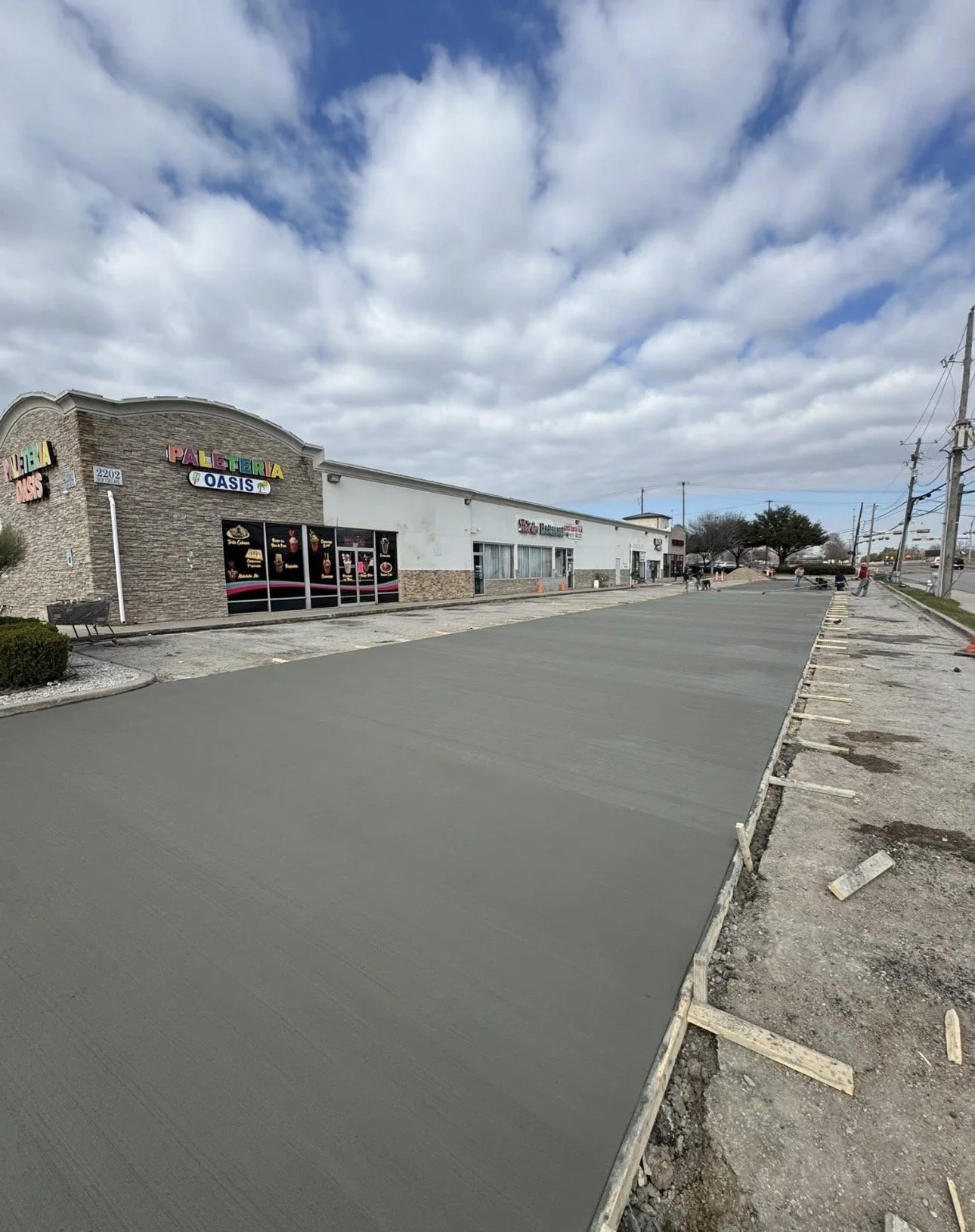 A commercial building with a sign that says 'Paleferia Oasis' in colorful letters, and a large, freshly poured concrete parking lot in front. The sky is cloudy with scattered clouds and there are utility poles and power lines visible along the street.