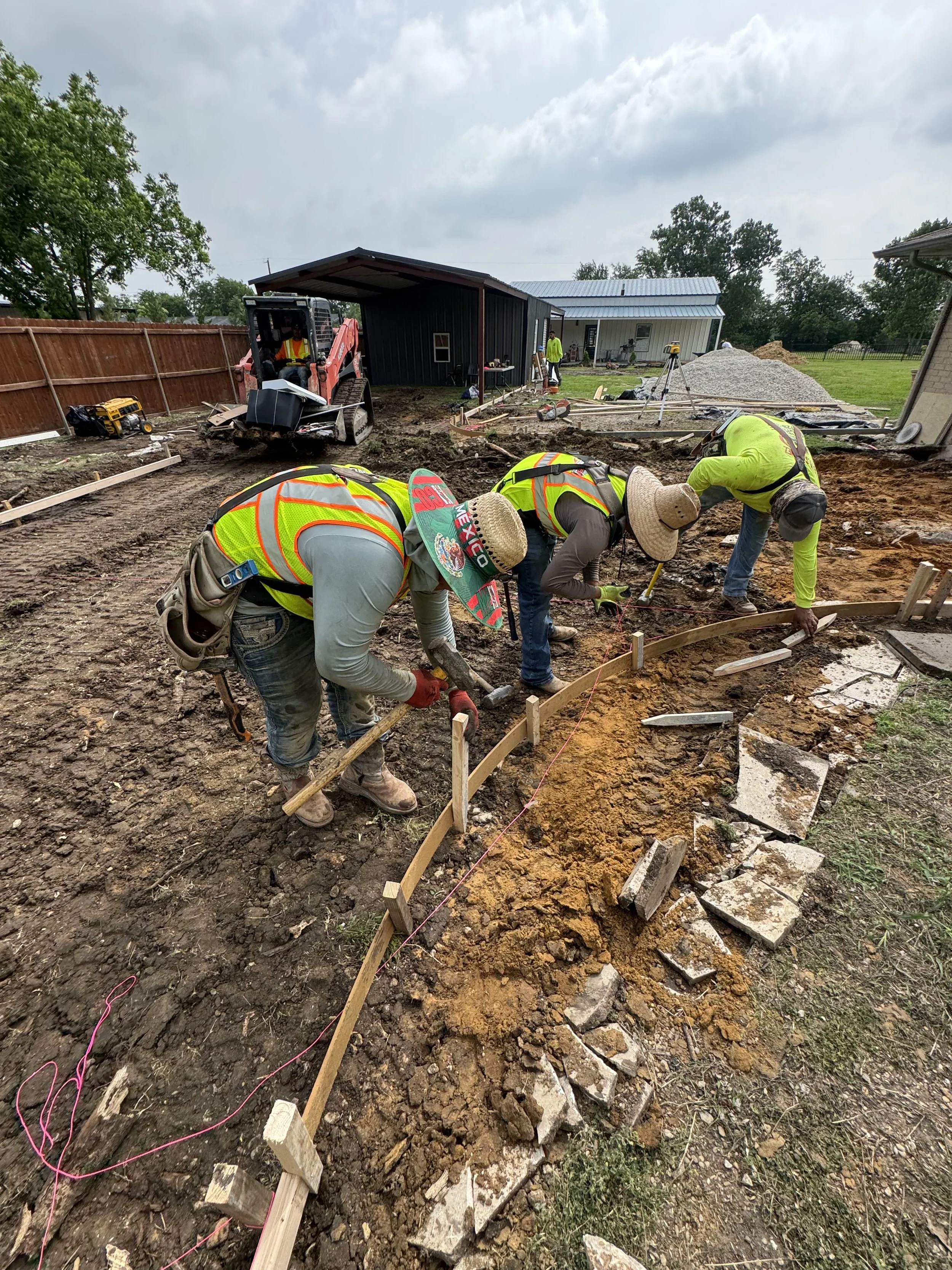Construction site with workers laying concrete, using tools and measuring equipment, with a house and a pile of gravel in the background.