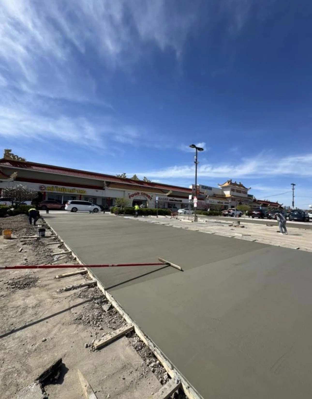 Construction workers leveling a concrete sidewalk in a shopping plaza parking lot under a partly cloudy sky.