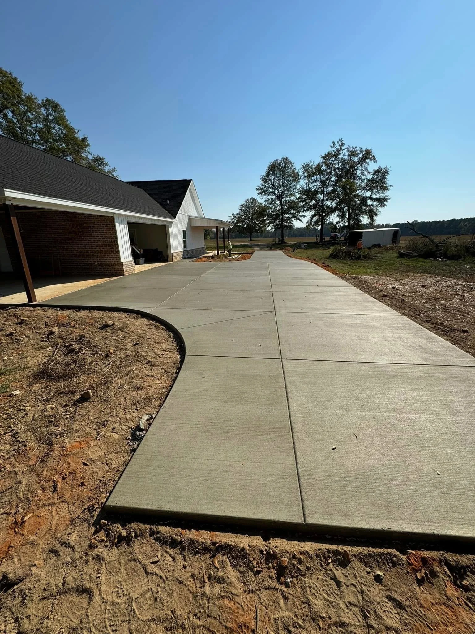 Newly poured concrete sidewalk in front of a house with a driveway, trees, and open field.
