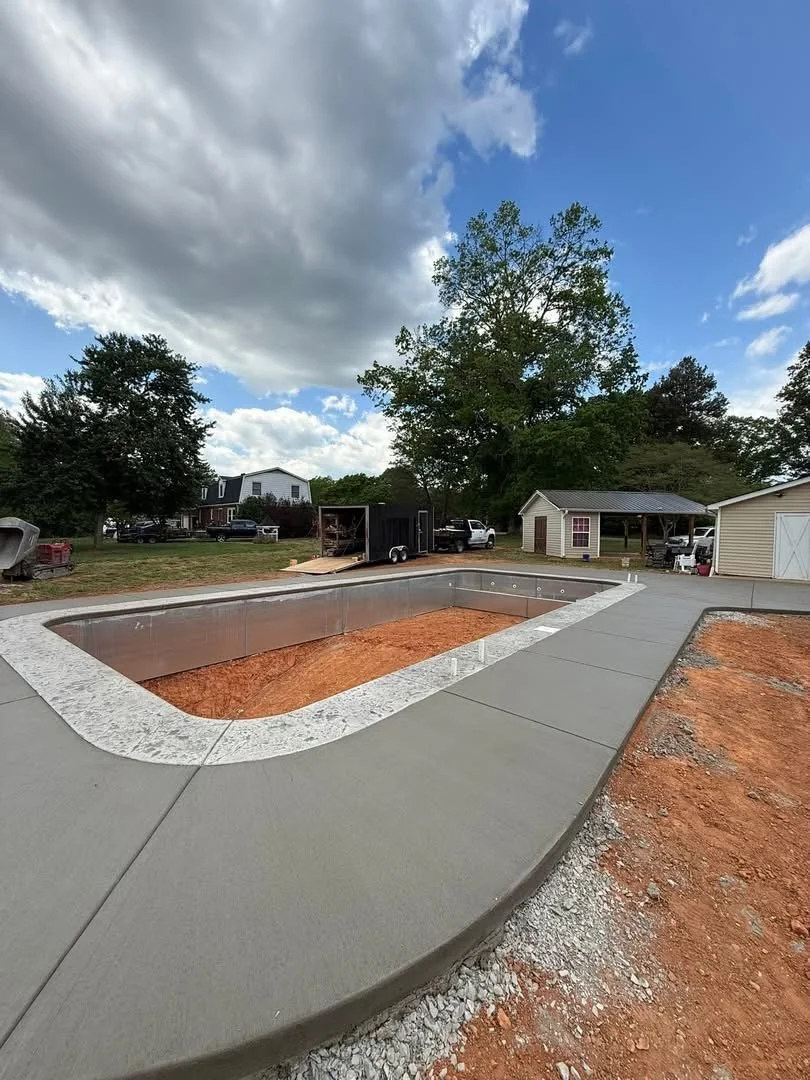 A backyard swimming pool under construction with a concrete deck around it, in a residential area with trees and houses in the background.