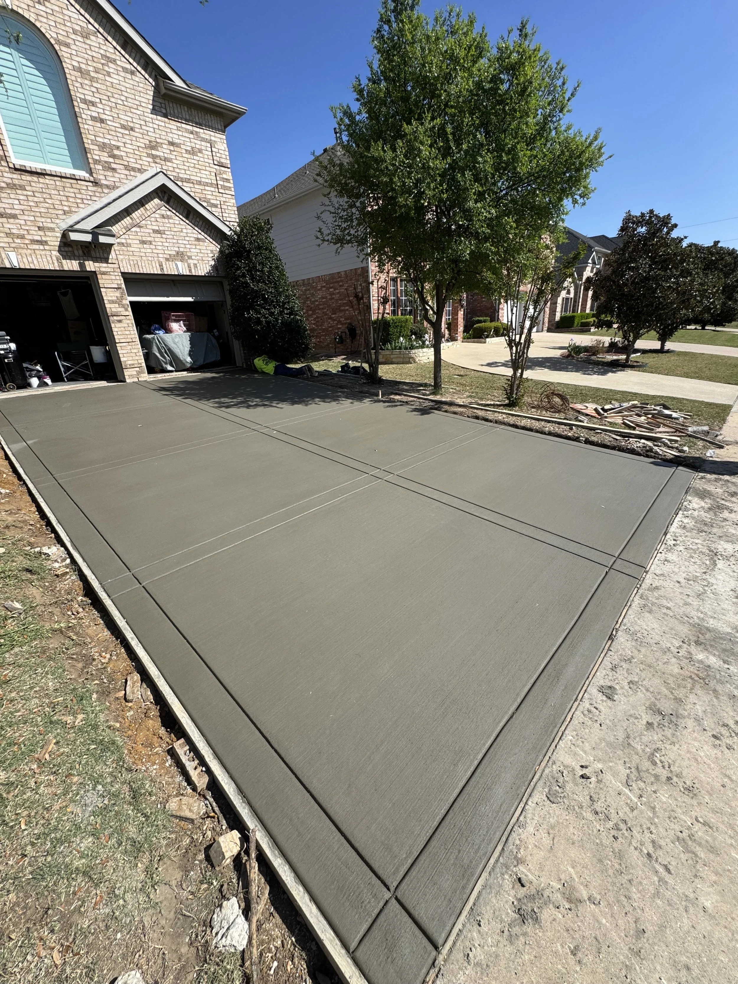 Newly poured concrete driveway in front of a house with a brick and siding exterior, trees, and landscaped bushes on a sunny day.