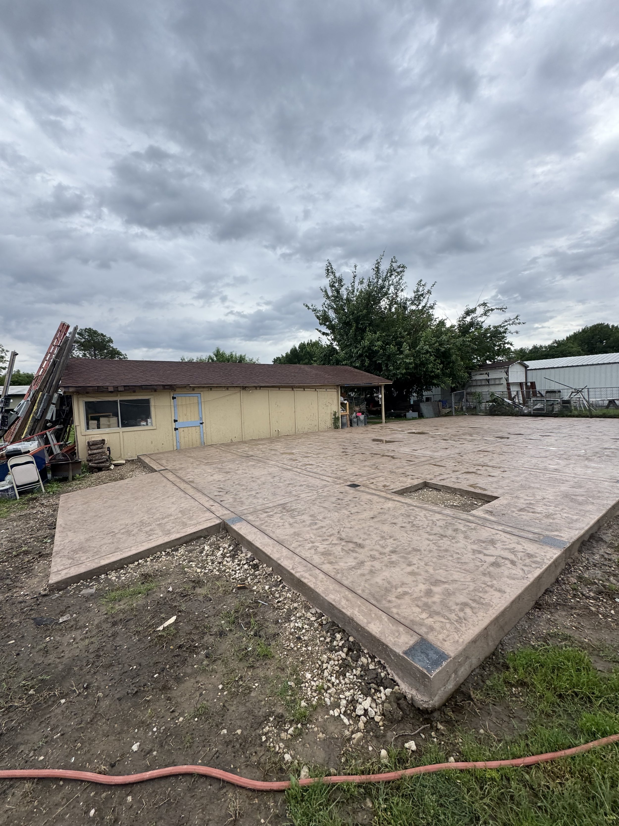 A backyard area with a small building, pallets, and construction materials. The ground has a concrete slab with some dirt and gravel. A red hose stretches across the dirt. The sky is overcast with gray clouds.