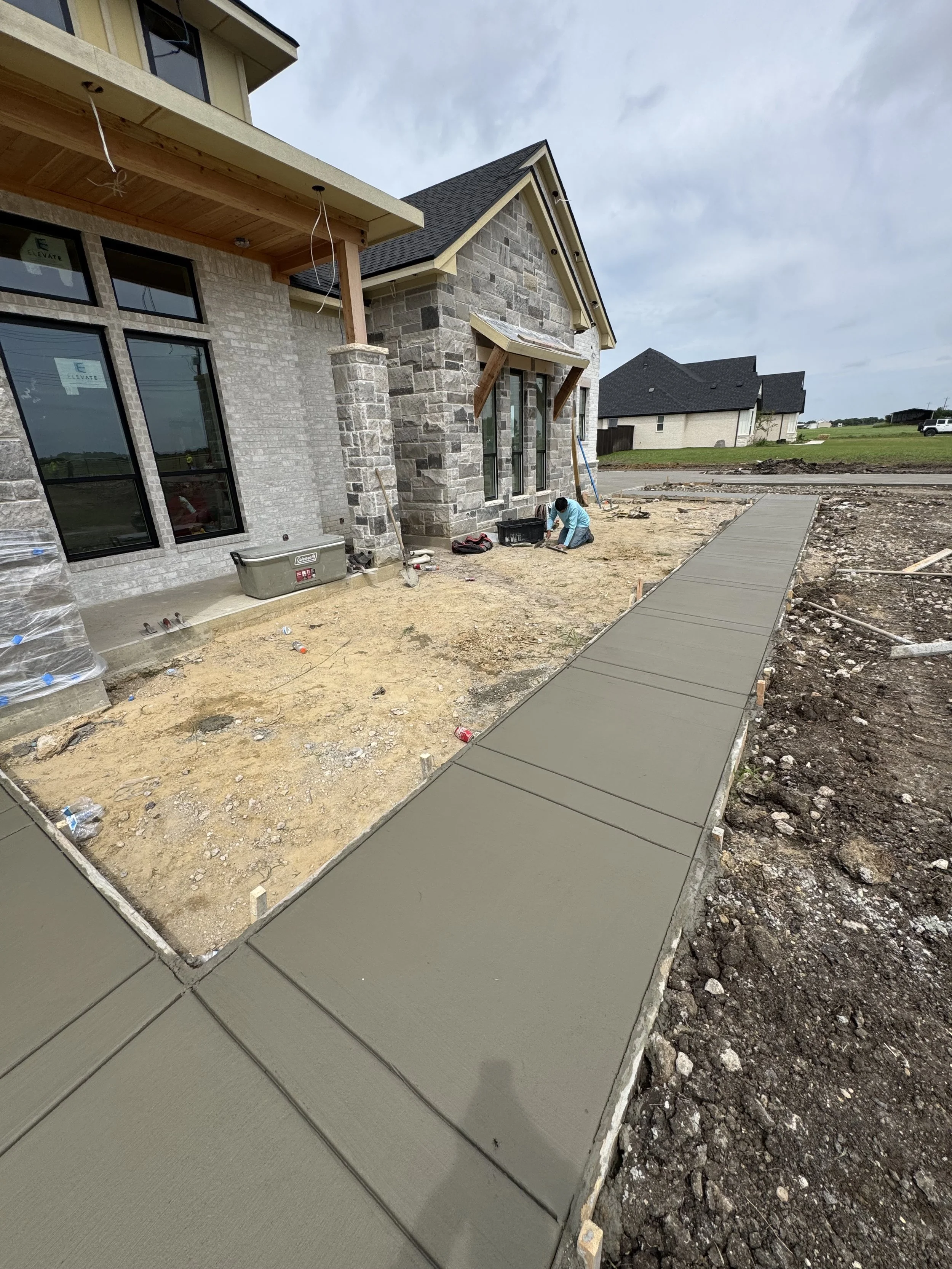 Construction workers building a concrete sidewalk in front of a house under construction, with a partially finished stone facade and a wooden porch roof.
