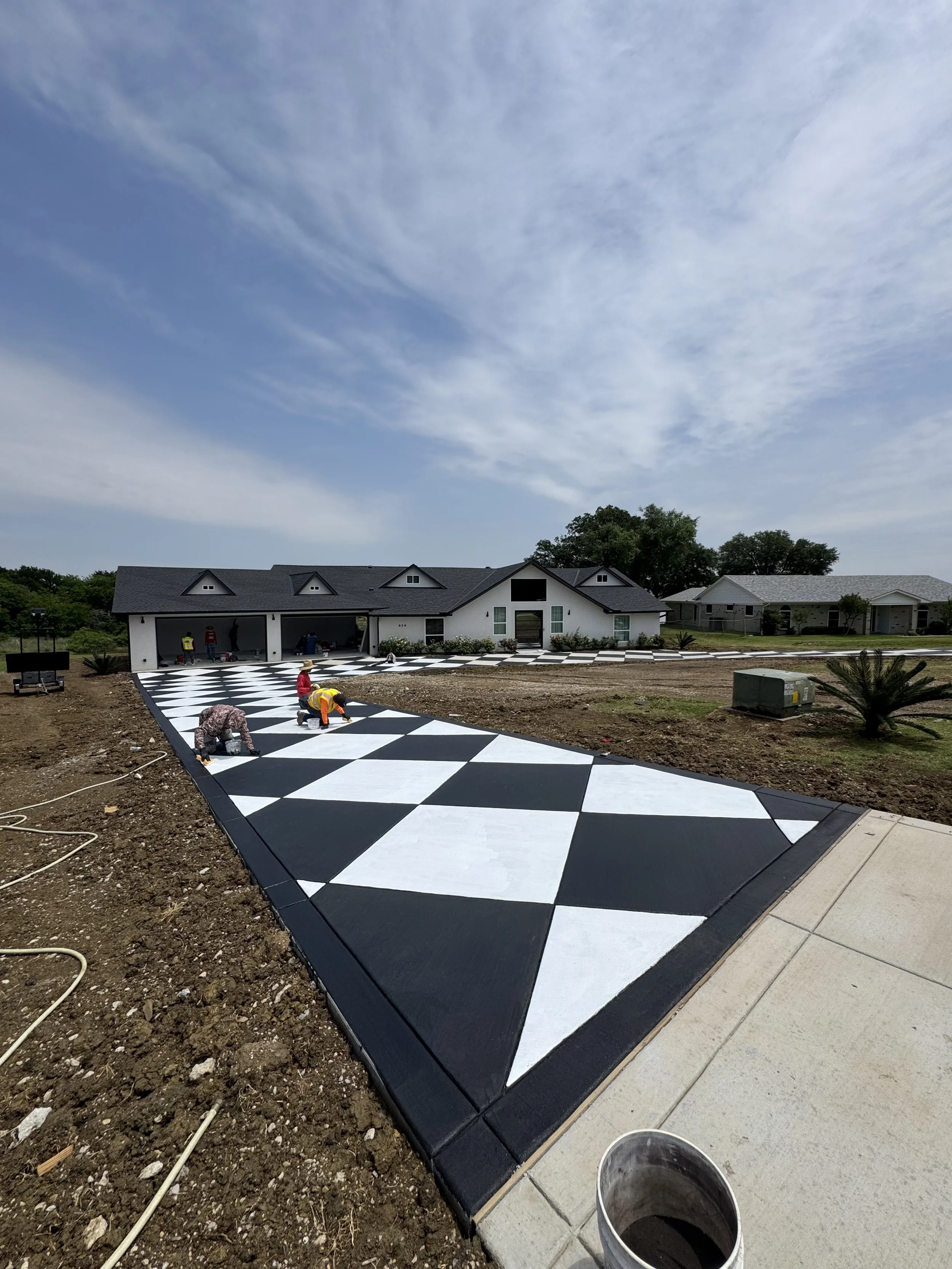Workers installing a black and white checkered pattern on a newly poured cement driveway in front of a house.