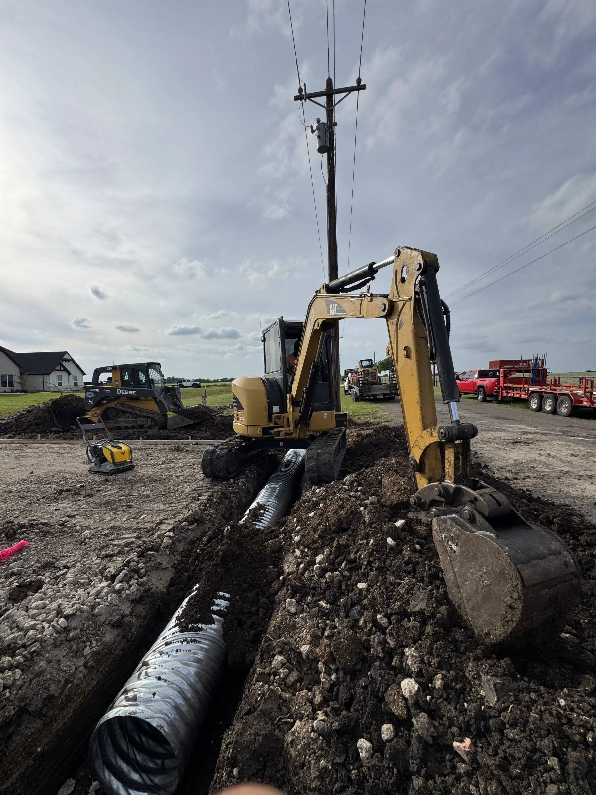 Construction site with excavator installing a large black pipe underground, other construction vehicles and equipment in the background, residential houses nearby, cloudy sky.